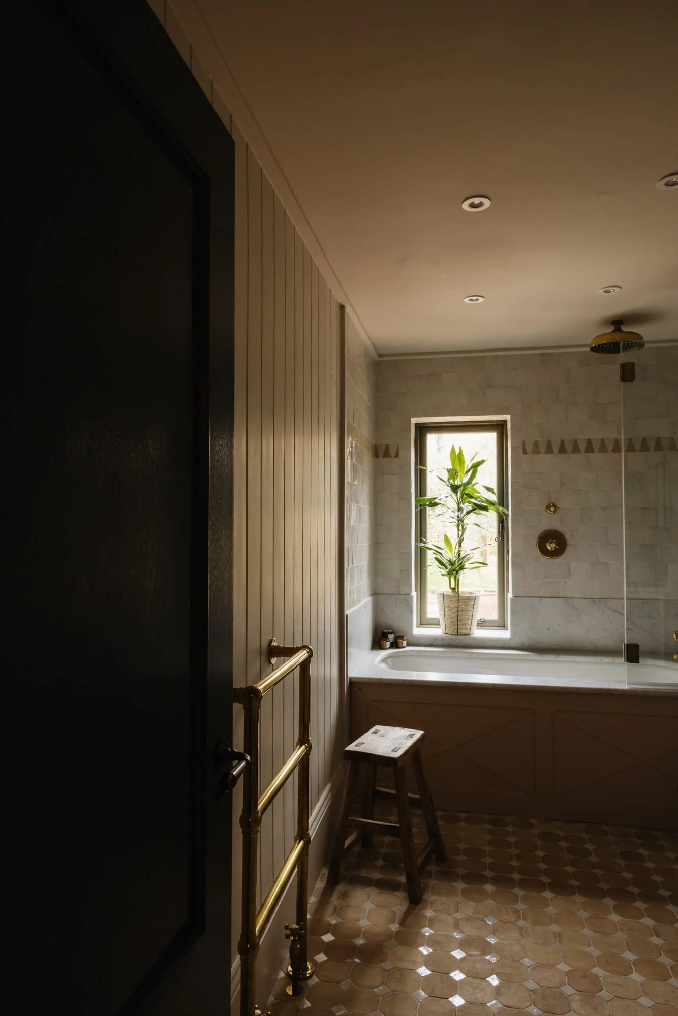 A cozy bathroom with a window, a potted plant, a bathtub, a small stool, and hexagonal tile flooring.