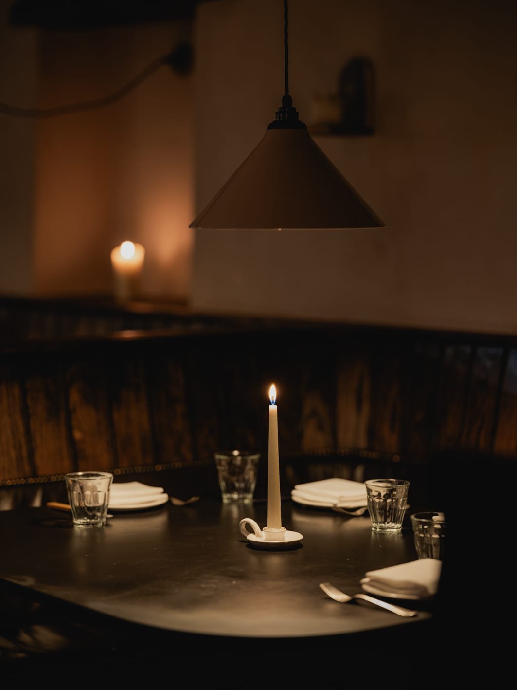 A dimly lit dining table set with a lit candle in the center, surrounded by glasses, plates, silverware, and a folded napkin, with a hanging light fixture above and candles in the background.