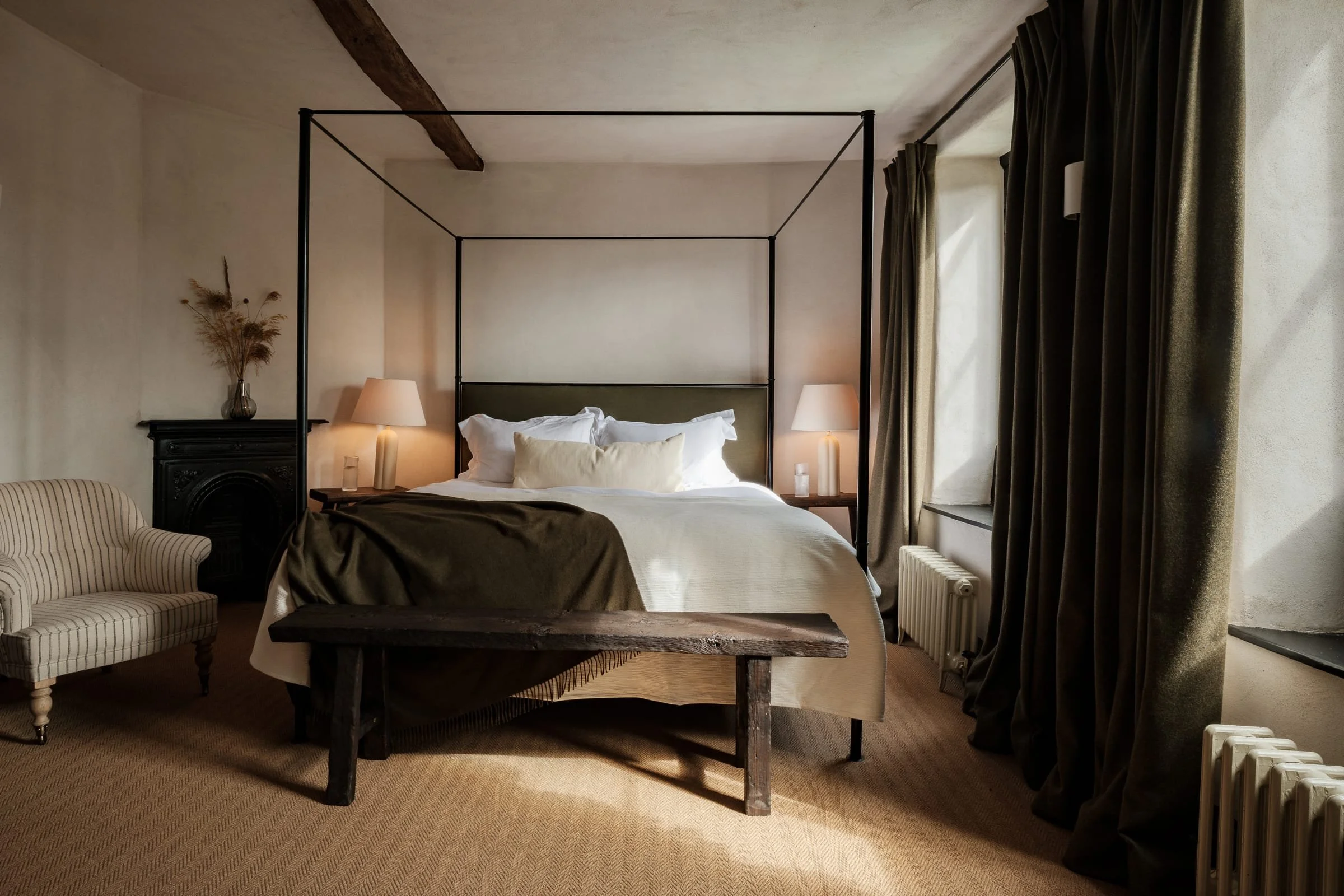 A bedroom with a four-poster bed, beige bedding, and pillows. There is a striped armchair, a black fireplace mantel with a vase of dried flowers, and two nightstands with lamps. Large windows with dark curtains are on the right.