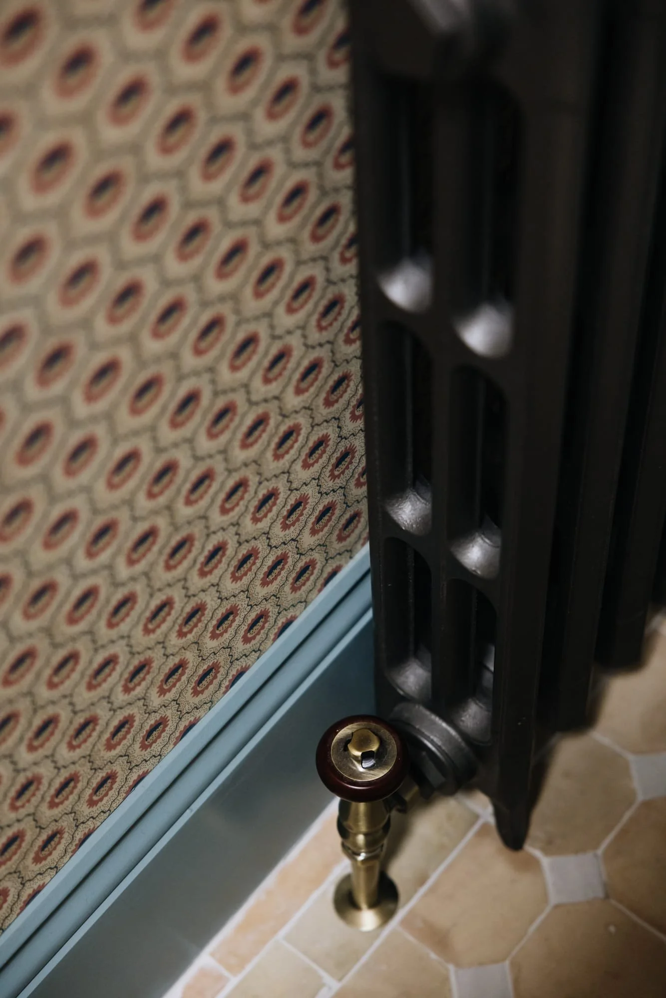 Close-up of a vintage black cast iron radiator with brass and wood valve, positioned next to a baseboard heater with patterned fabric covering, on a tiled floor.