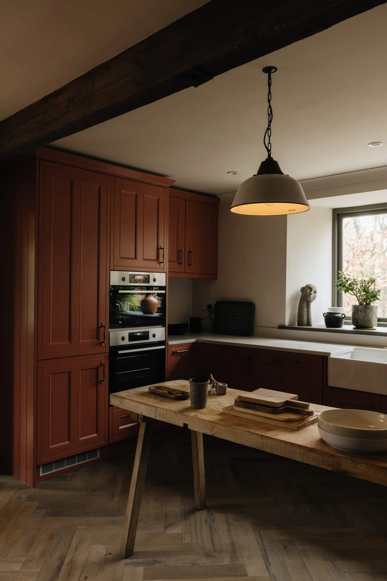 A cozy kitchen with red wooden cabinets, a white farmhouse sink, a window with potted plants, and a wooden table with bowls and cutting boards, illuminated by a hanging pendant light.