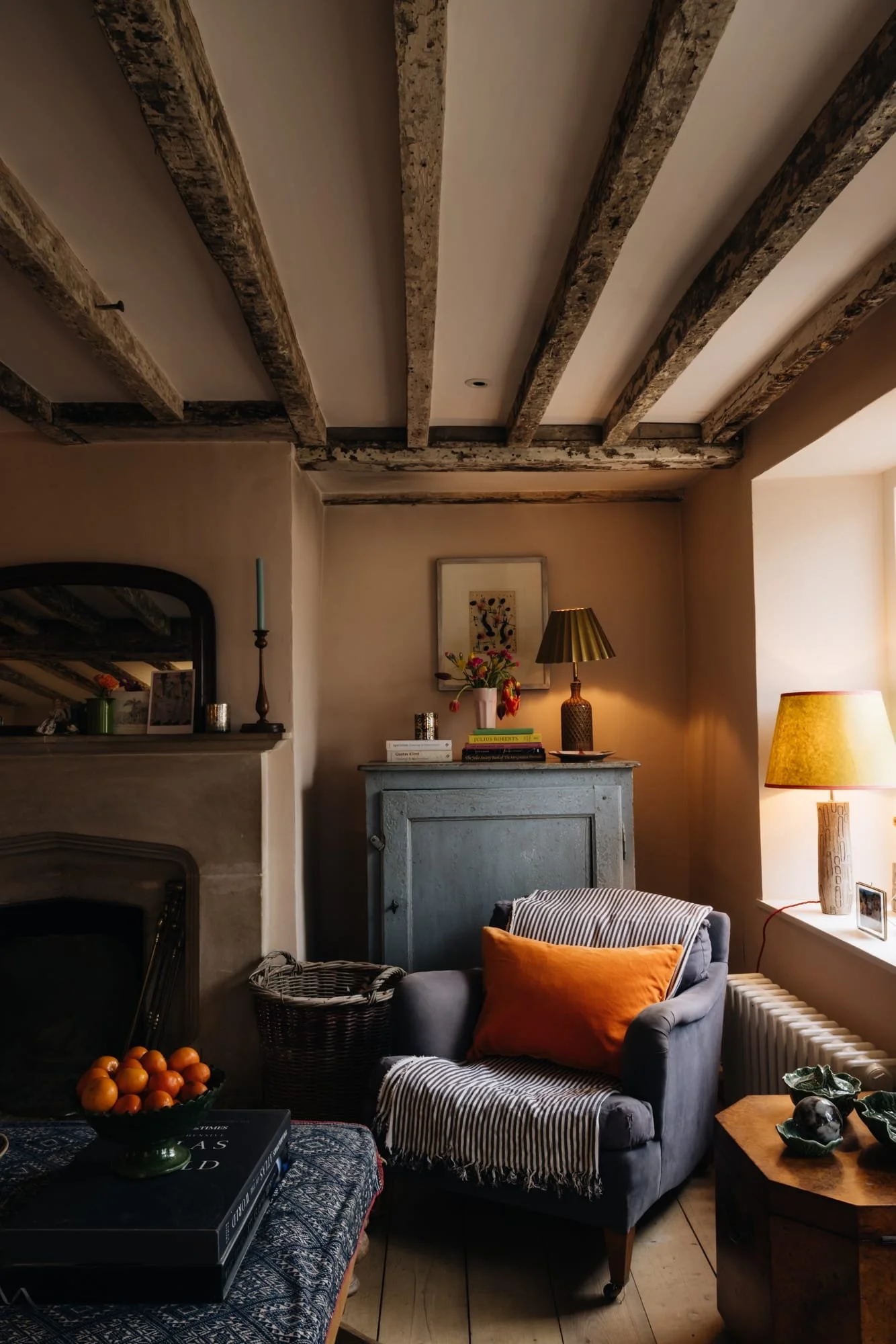 Cozy living room with exposed wooden ceiling beams, a gray armchair with an orange pillow, a blue cabinet with decorative items, and warm lighting from a window and lamps.