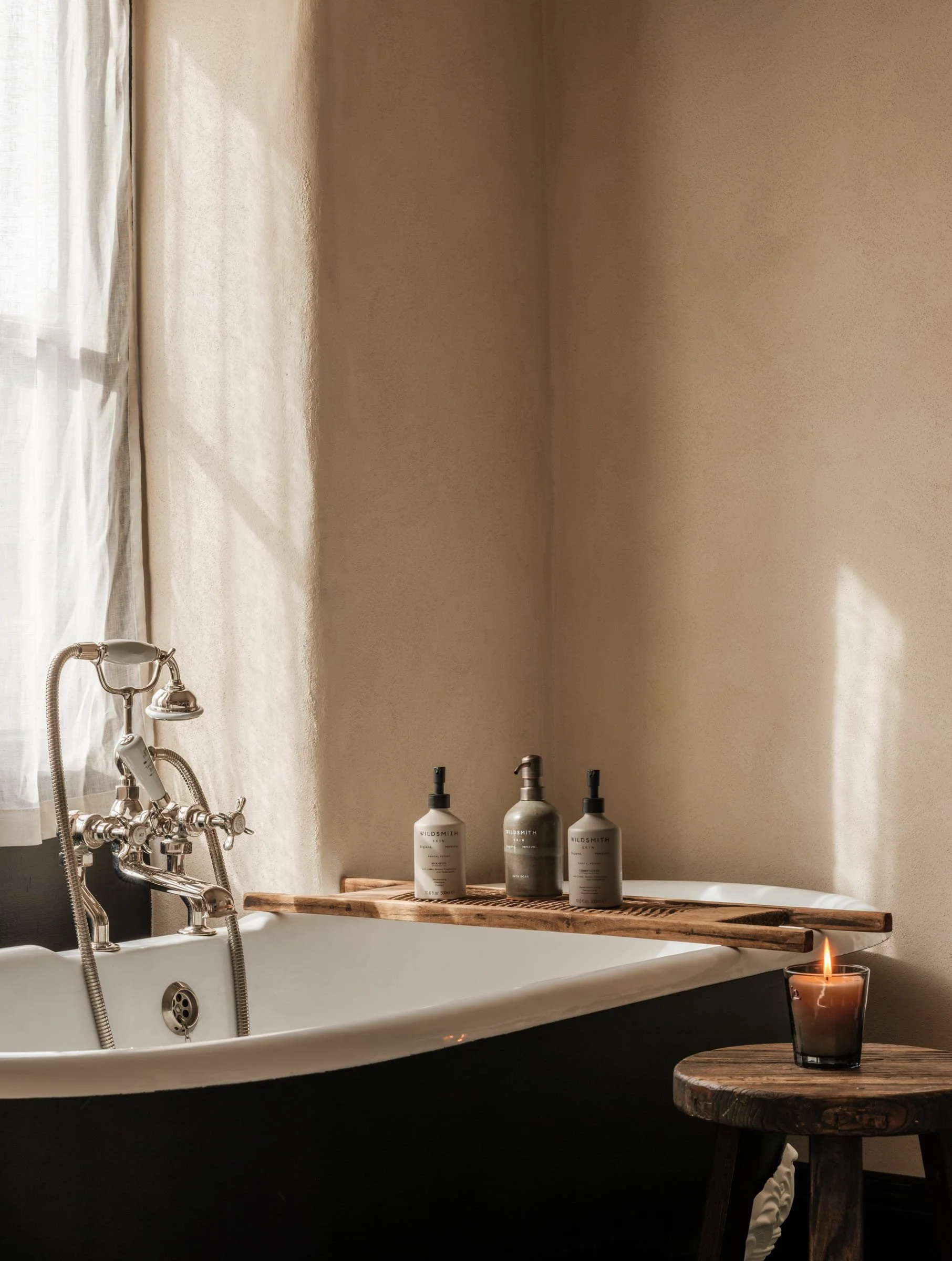 A vintage-style bathroom with a clawfoot tub and a lit candle on a small wooden stool, with a wooden tray holding bathroom bottles on the edge of the tub, near a window with sheer curtains.