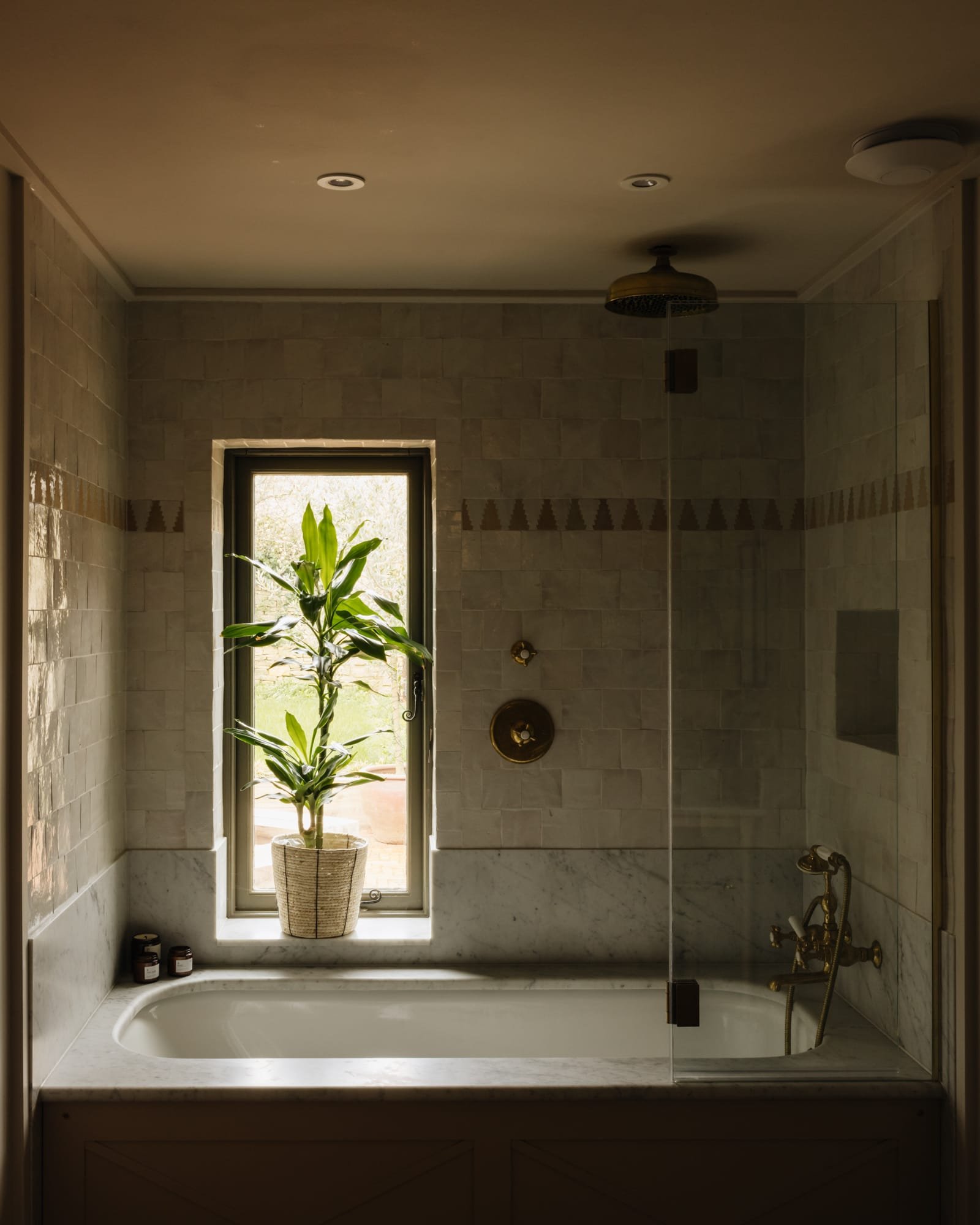 A bathroom with a built-in bathtub, a large window with a potted plant, and beige tiled walls.