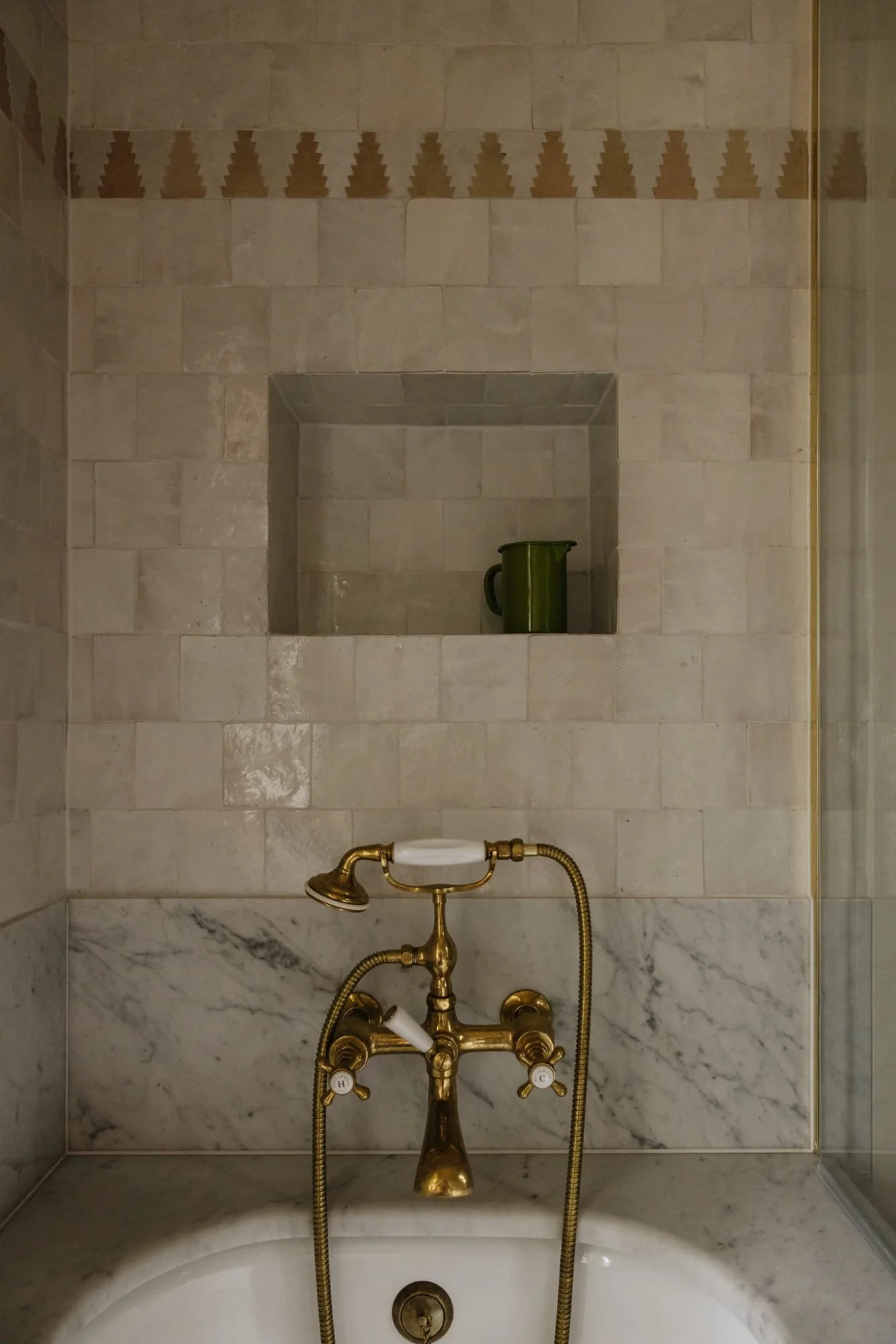 A bathroom with a bathtub, featuring vintage-style gold fixtures, a tiled wall with a built-in niche holding a green pitcher, and a marble backsplash above the tub.