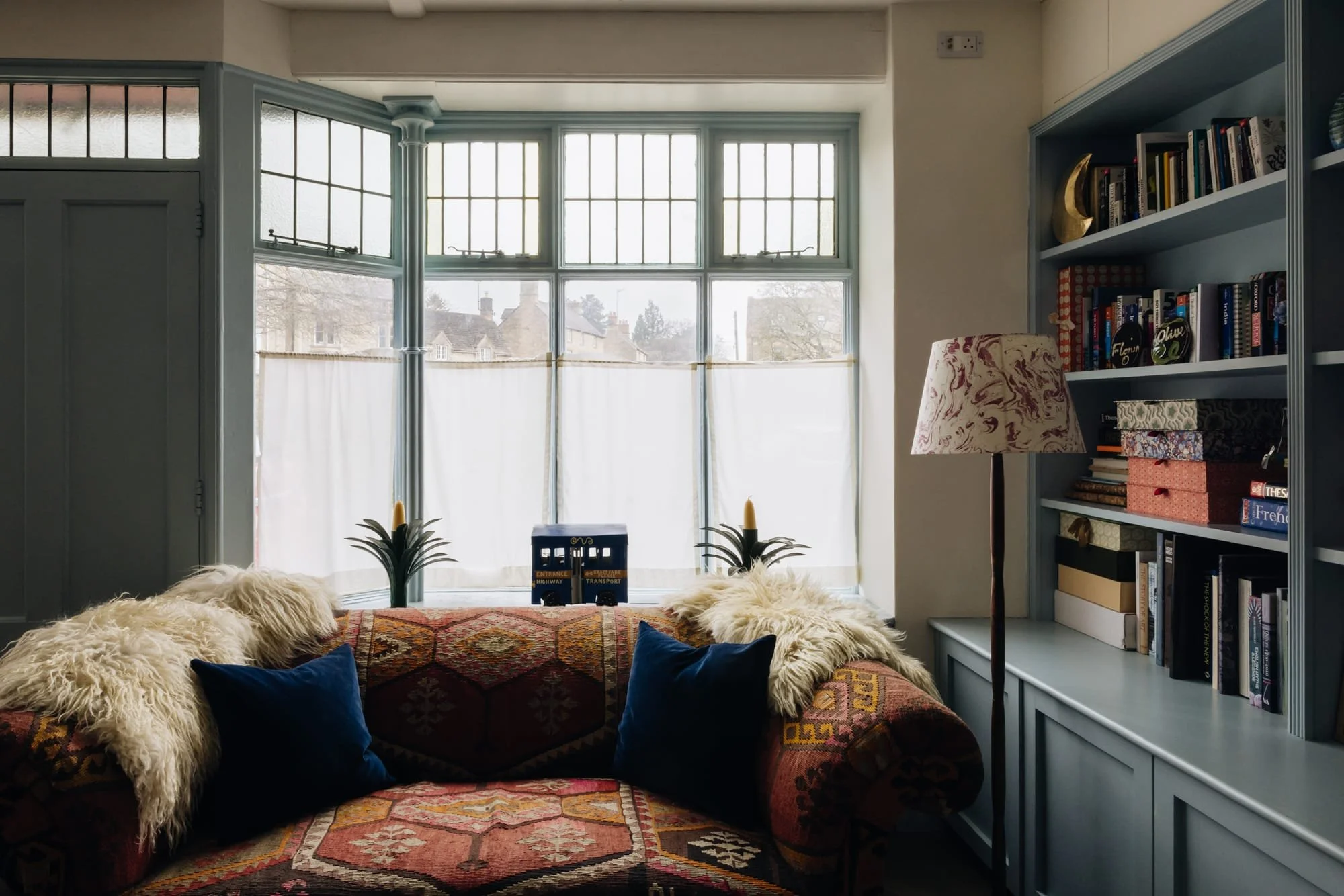 Living room with large bay window, blue shelving unit filled with books, a floral patterned floor lamp, and a vintage sofa with colorful upholstery and blue pillows, decorated with faux fur throws and two small artificial plants, with sunlight coming