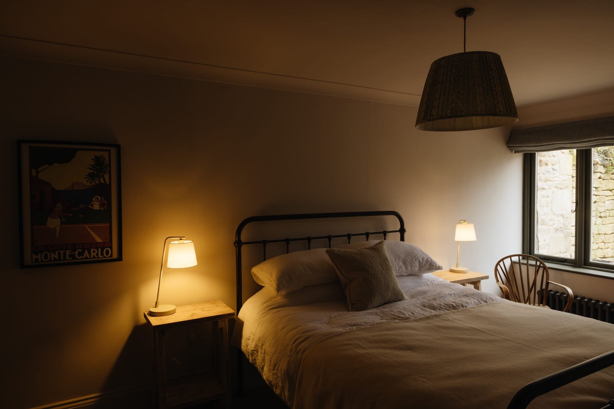 A cozy bedroom with a bed, two small bedside tables and lamps, a poster on the wall, a window with a view of a stone wall outside, and a wooden chair beside the window. The room has warm lighting.