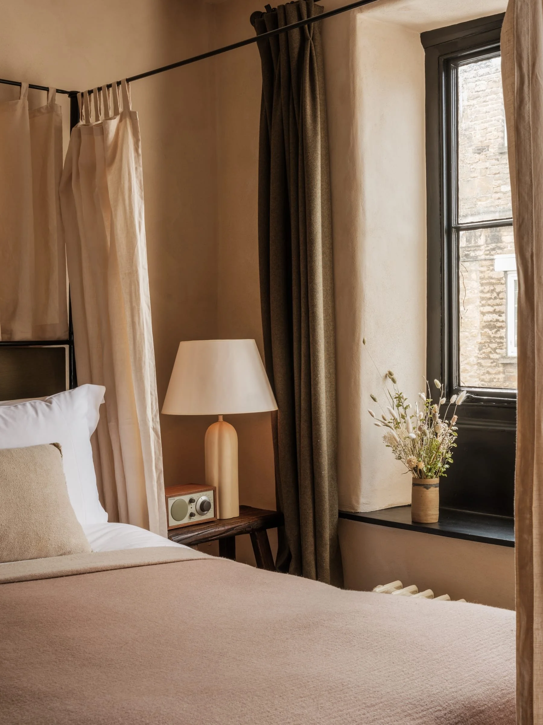 A cozy bedroom corner with a bed, beige bedding, a white pillow, a nightstand with a lamp, a small radio, a flower vase with white flowers, and bamboo and dark curtain window coverings.
