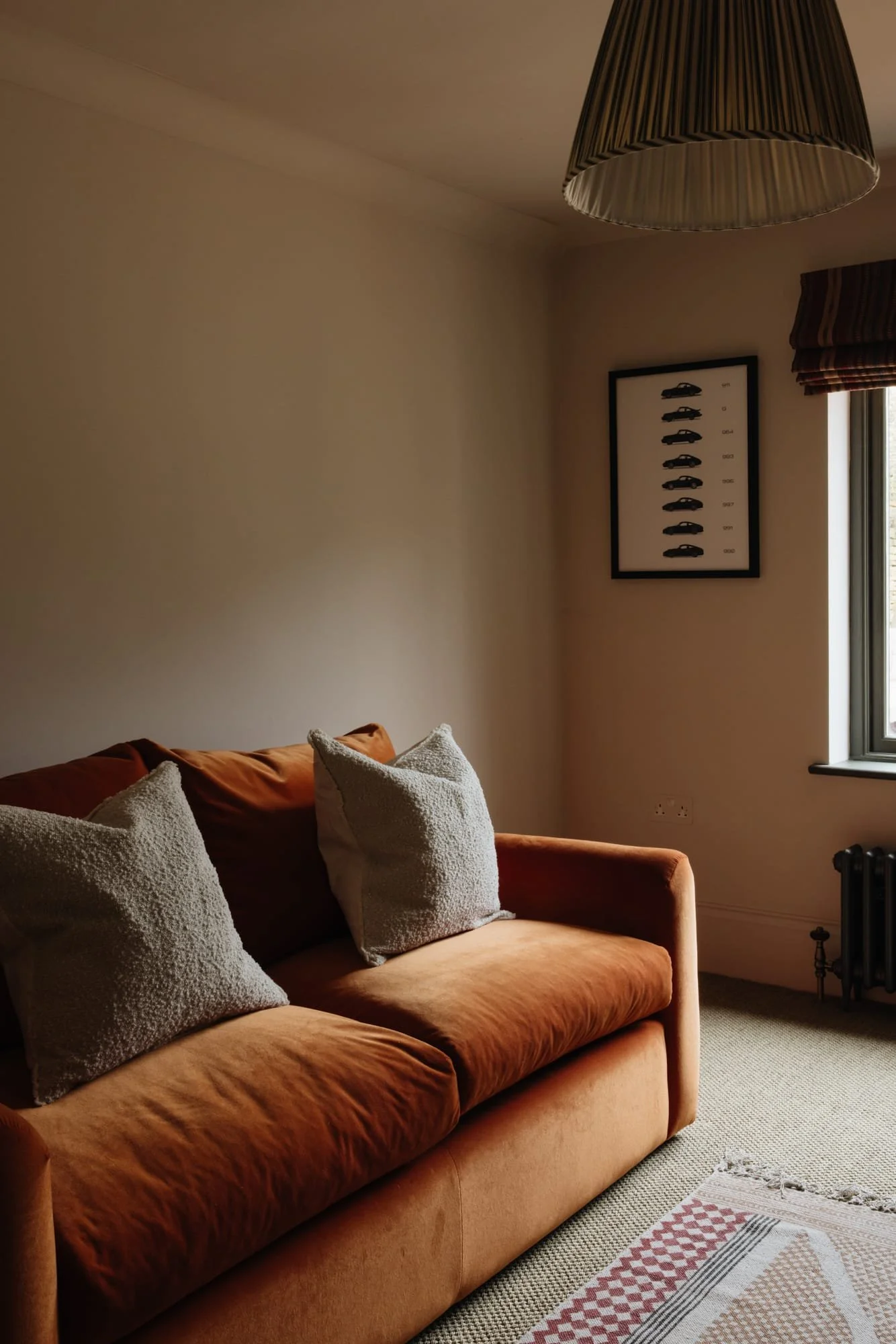 A cozy living room with an orange velvet sofa and beige throw pillows, a window with striped curtains, a framed car chart on the wall, a ceiling light, and a patterned rug on a beige carpet.