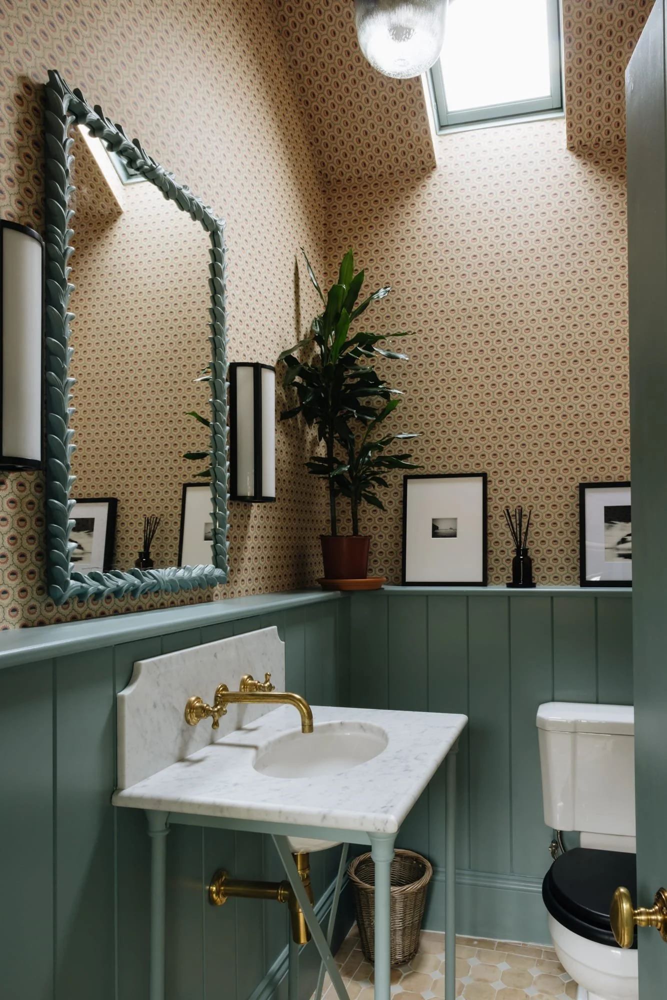 Bathroom with patterned wallpaper, green wainscoting, gold fixtures, rectangular mirror with twisted frame, potted plant, framed black-and-white photographs, wall-mounted light fixtures, skylight, and a small white marble sink.