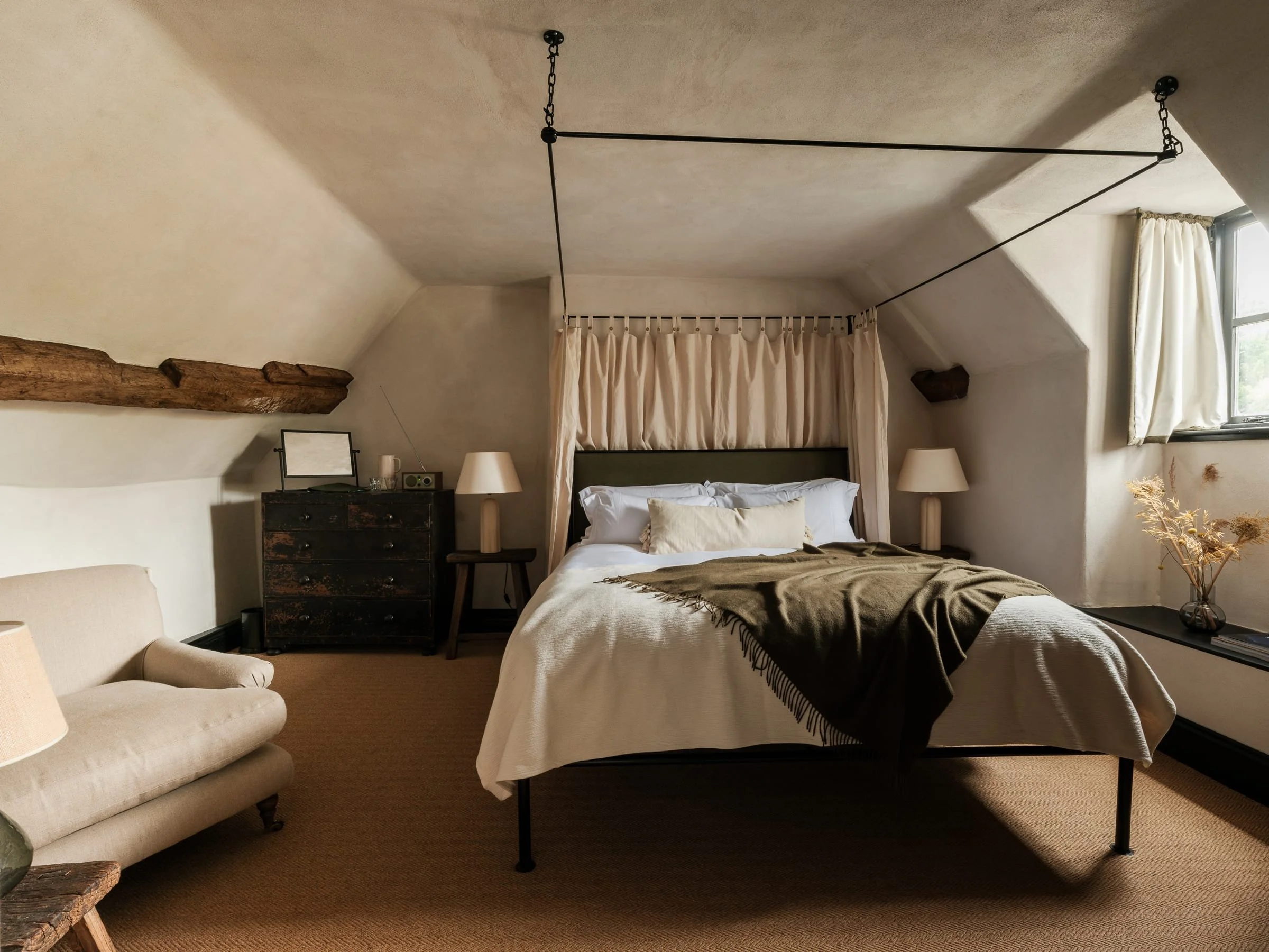 A cozy bedroom with a sloped ceiling, a bed with white bedding and a dark throw blanket, two bedside lamps, a black dresser, a beige armchair, window with curtains, and decorative dried plants.
