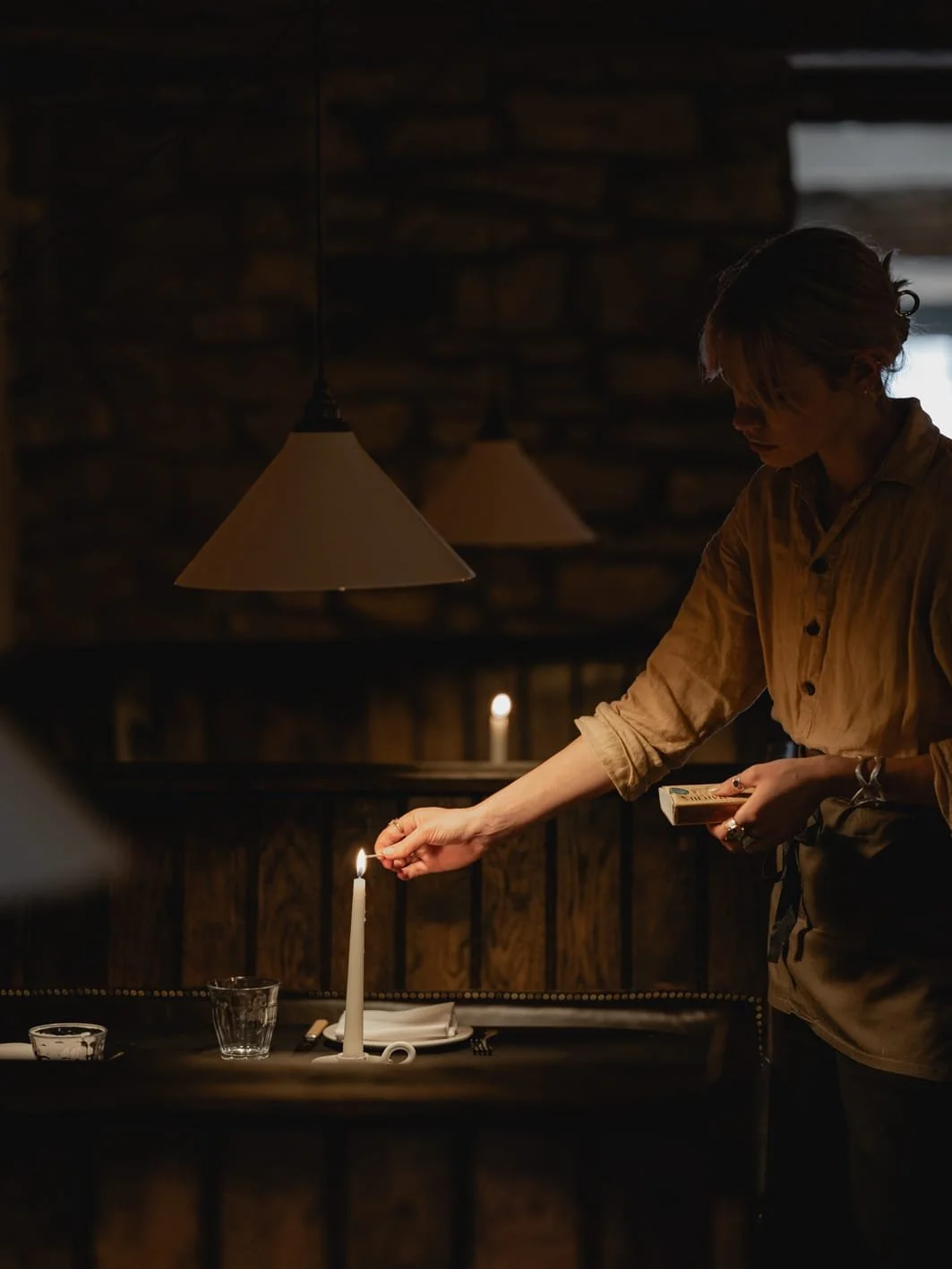 A woman lighting a candle on a dining table in a dimly lit room, with a stone wall in the background and hanging lamps above.
