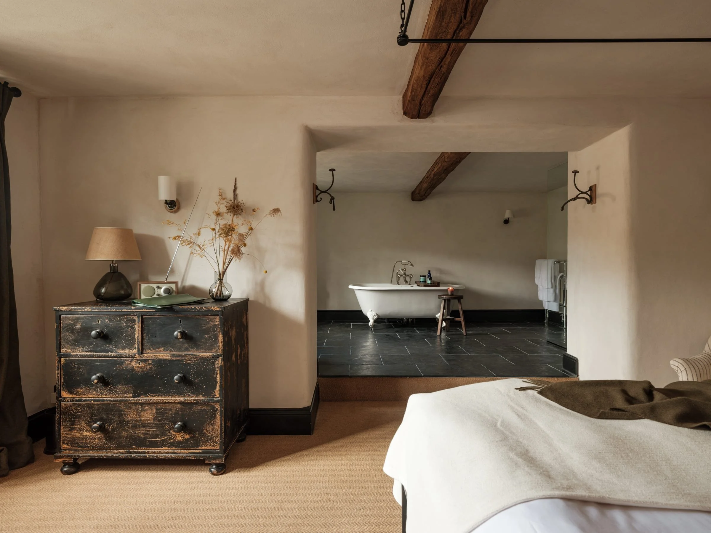 A rustic bedroom with a vintage black dresser topped with a lamp, a vase of dried flowers, and a radio. A bed with white linens is partially visible in the foreground, and a small bathroom with a clawfoot bathtub, black tile flooring, and a towel rac