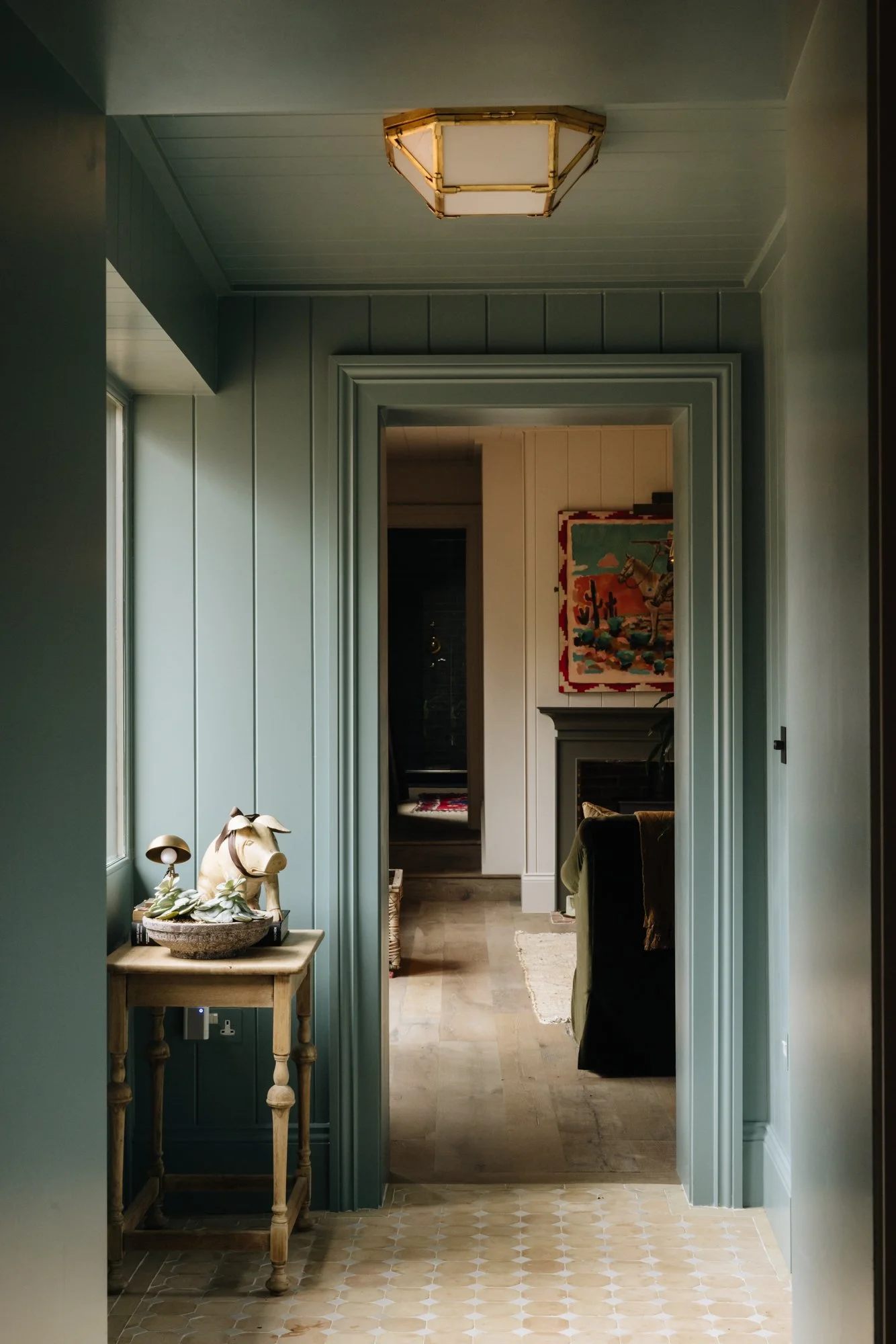 View of a hallway with green walls, a wooden table with decorative items, leading to a living room with artwork and a fireplace.