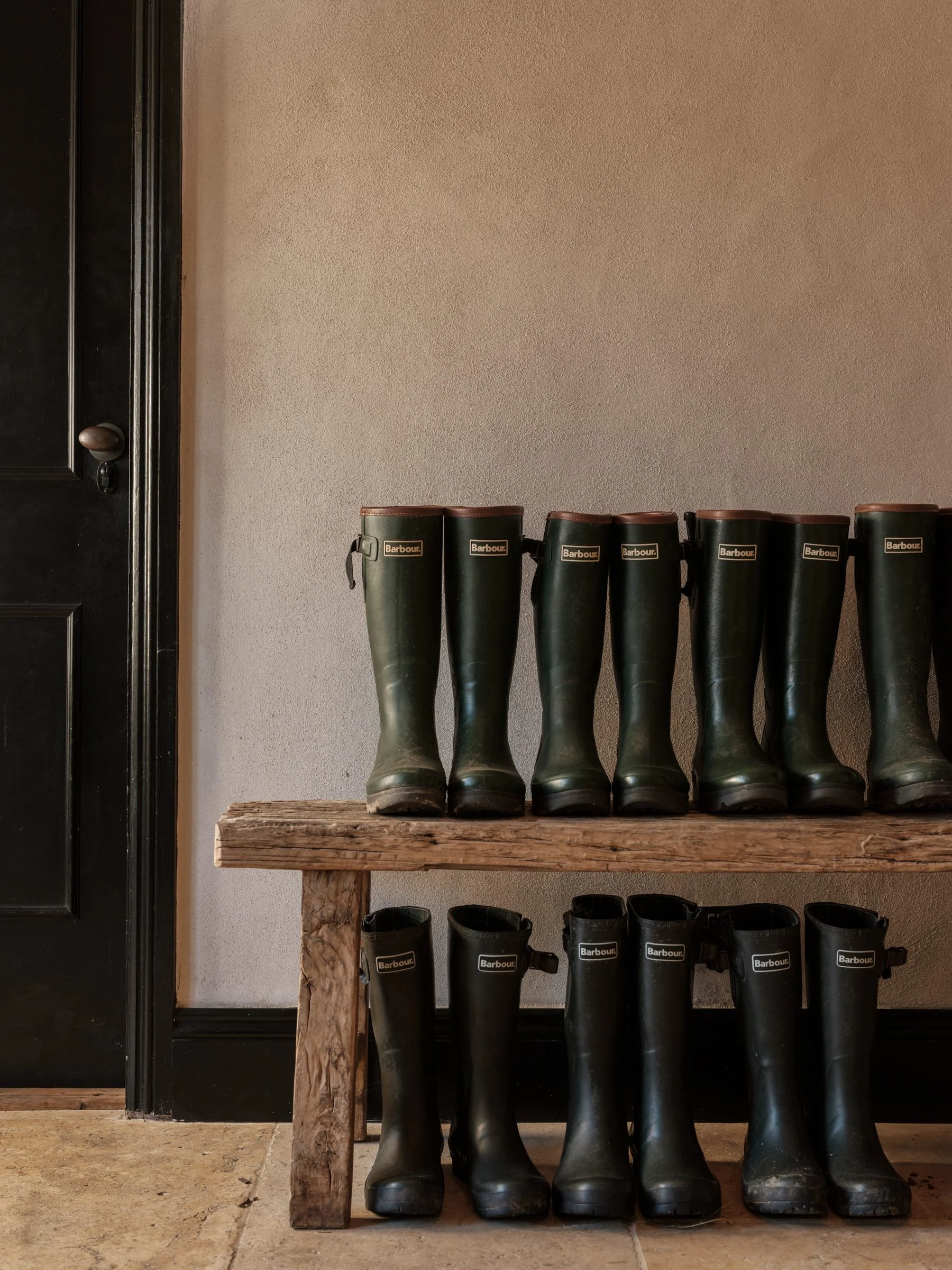 A pair of green rubber boots on top of a wooden bench and another pair of black rubber boots underneath on a tiled floor, with a beige wall and black door in the background.