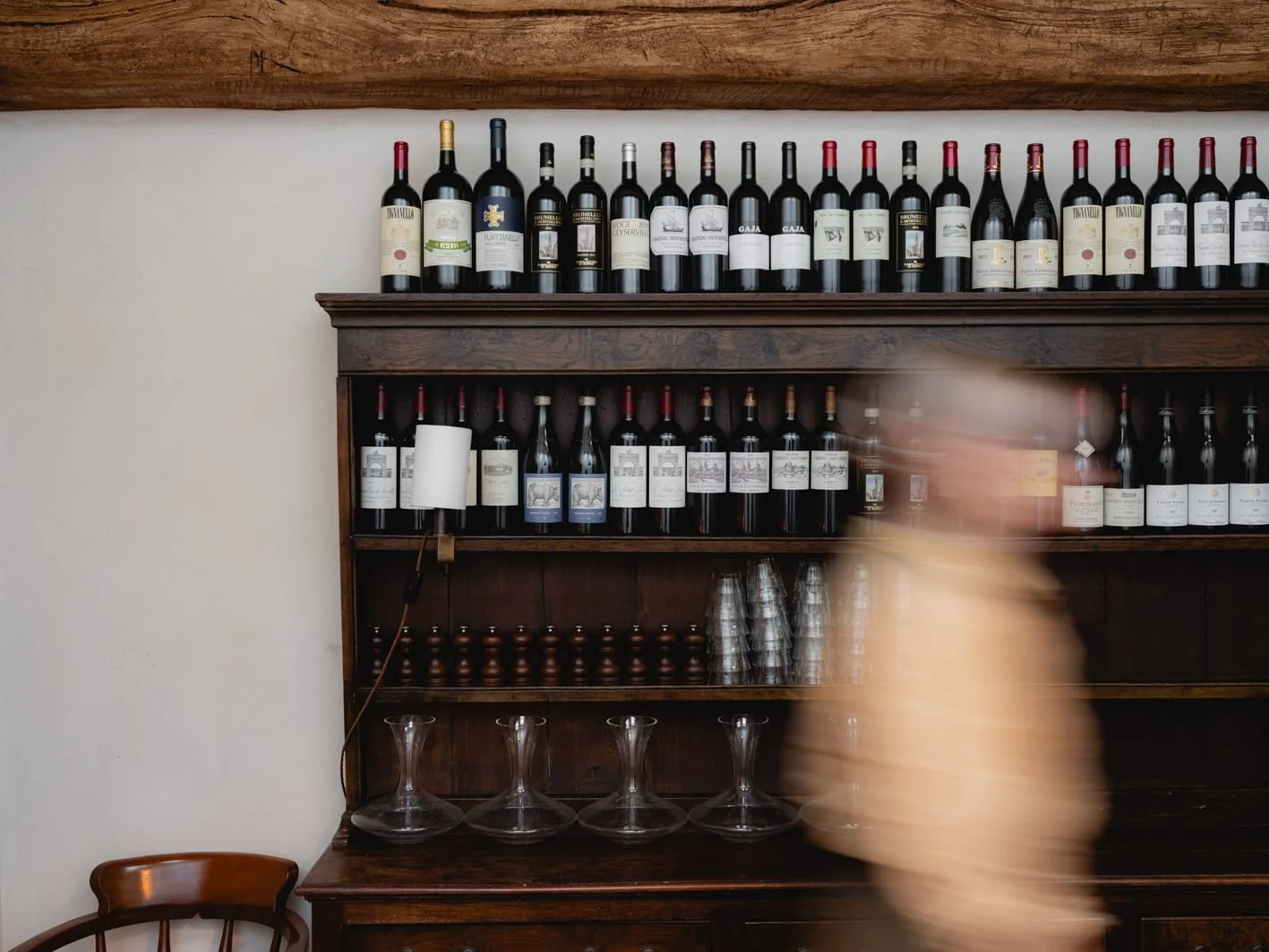 Wall-mounted wooden wine rack filled with bottles of red wine, under which are glasses and wine accessories, with a person blurred in motion walking past.