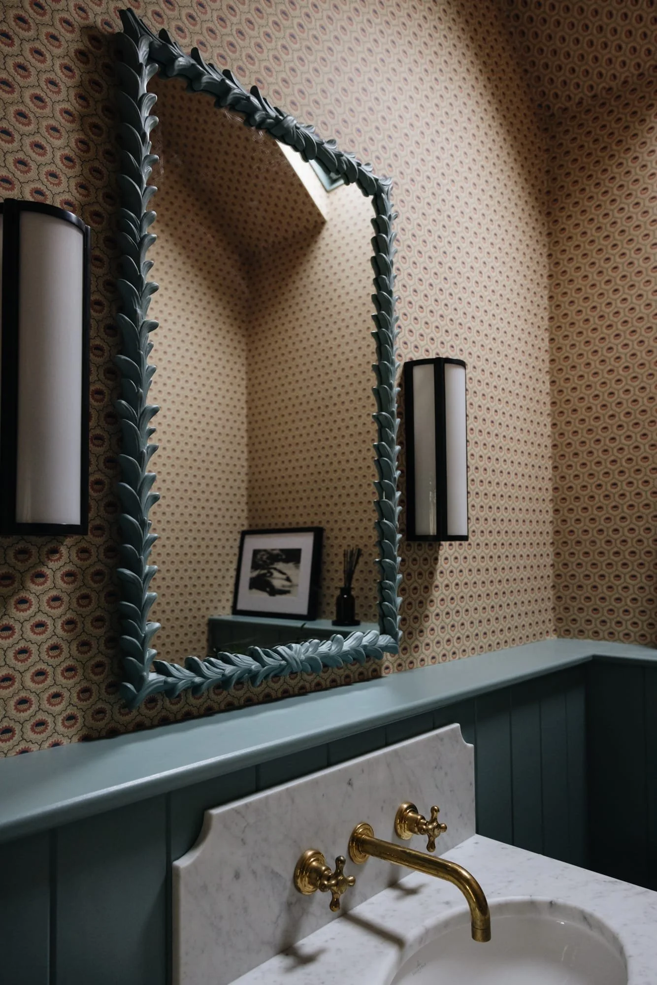 A bathroom with a decorative mirror, wallpaper with a geometric pattern, wall sconces, a framed black-and-white photo, and a marble-topped sink with gold fixtures.