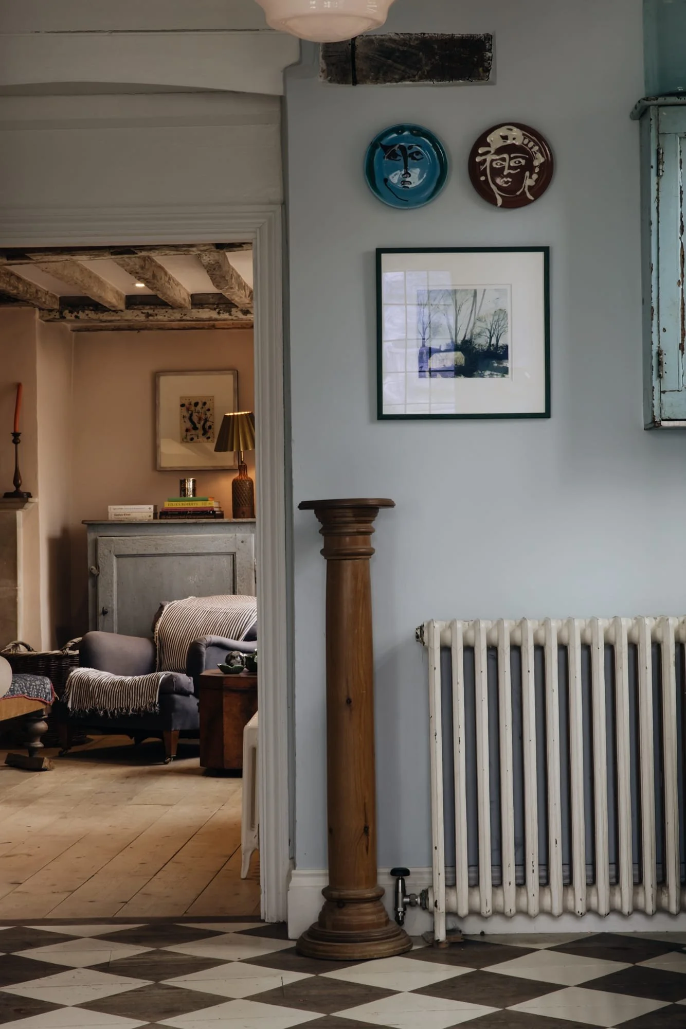 Interior view of a cozy living room with a light blue wall, framed artwork, two decorative plates on the wall, a wooden column, and a white radiator. Part of another room with a gray armchair, floor lamp, and decorative items is visible through an op