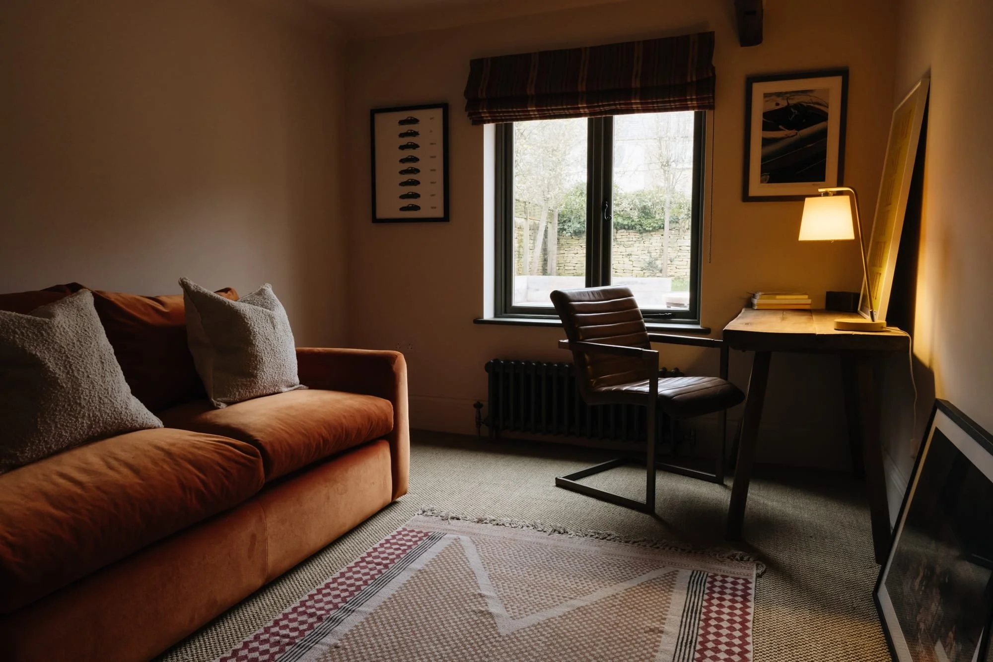 A cozy living room with a brown sofa, white pillows, a wooden desk with a lamp and books, a leather chair, a window with blinds, a framed art print, and a patterned rug.