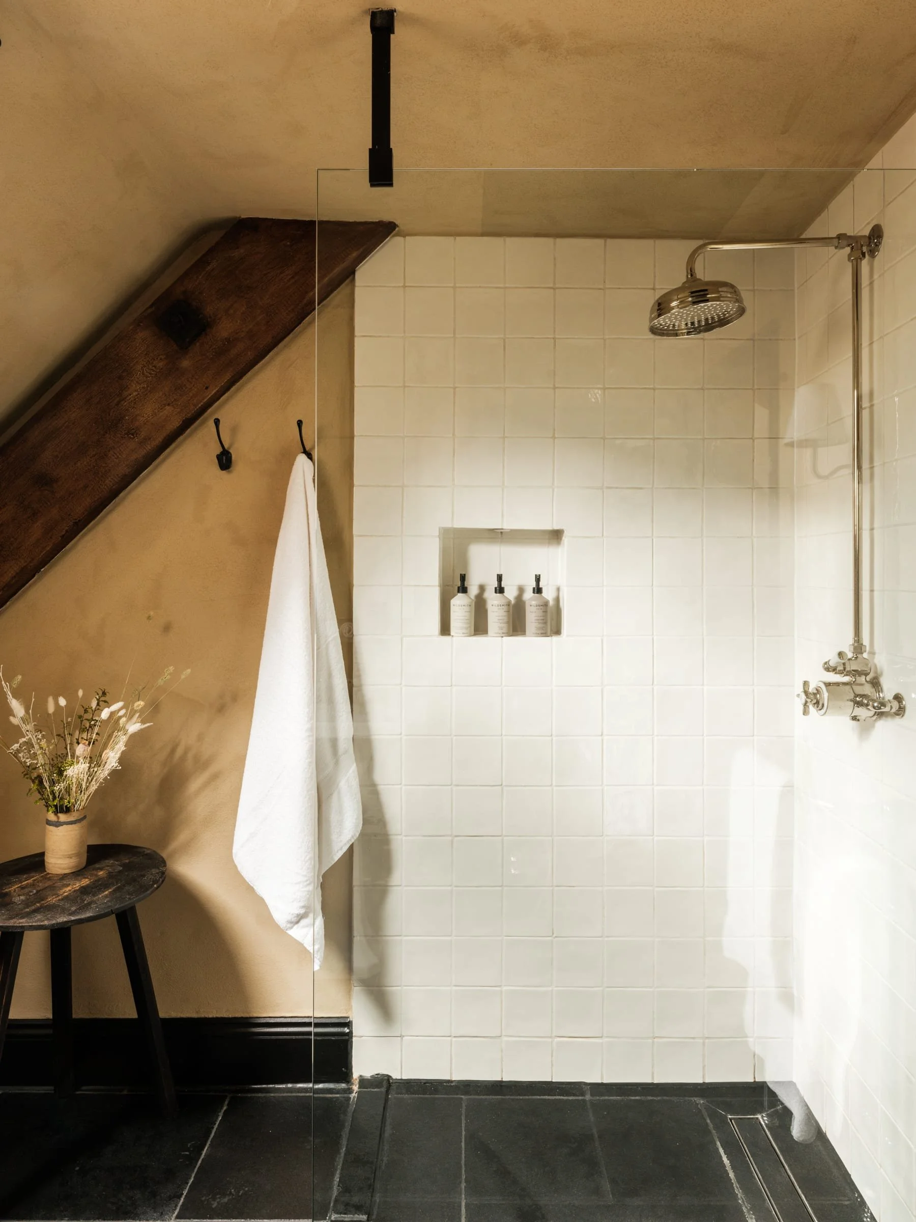 Modern shower area with white tile walls, a rain shower head, a nook with three bottles, and a small wooden side table with a vase of dried flowers, all under a sloped ceiling with a wooden beam.