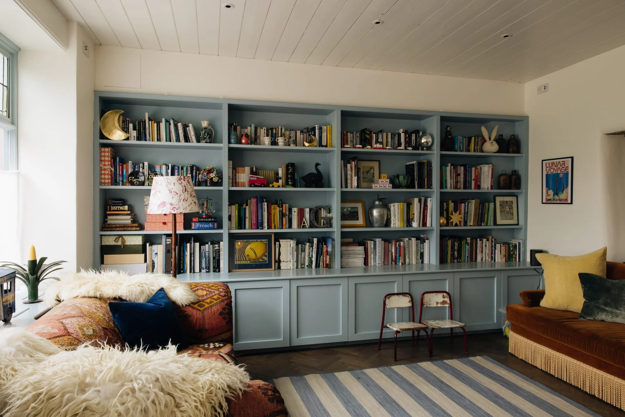 A cozy living room with a large, light blue bookshelf filled with books, decorative items, and framed pictures. To the left, there is a window with two chairs in front of it, a floral lamp, and a plush blanket on a colorful armchair. To the right, a 