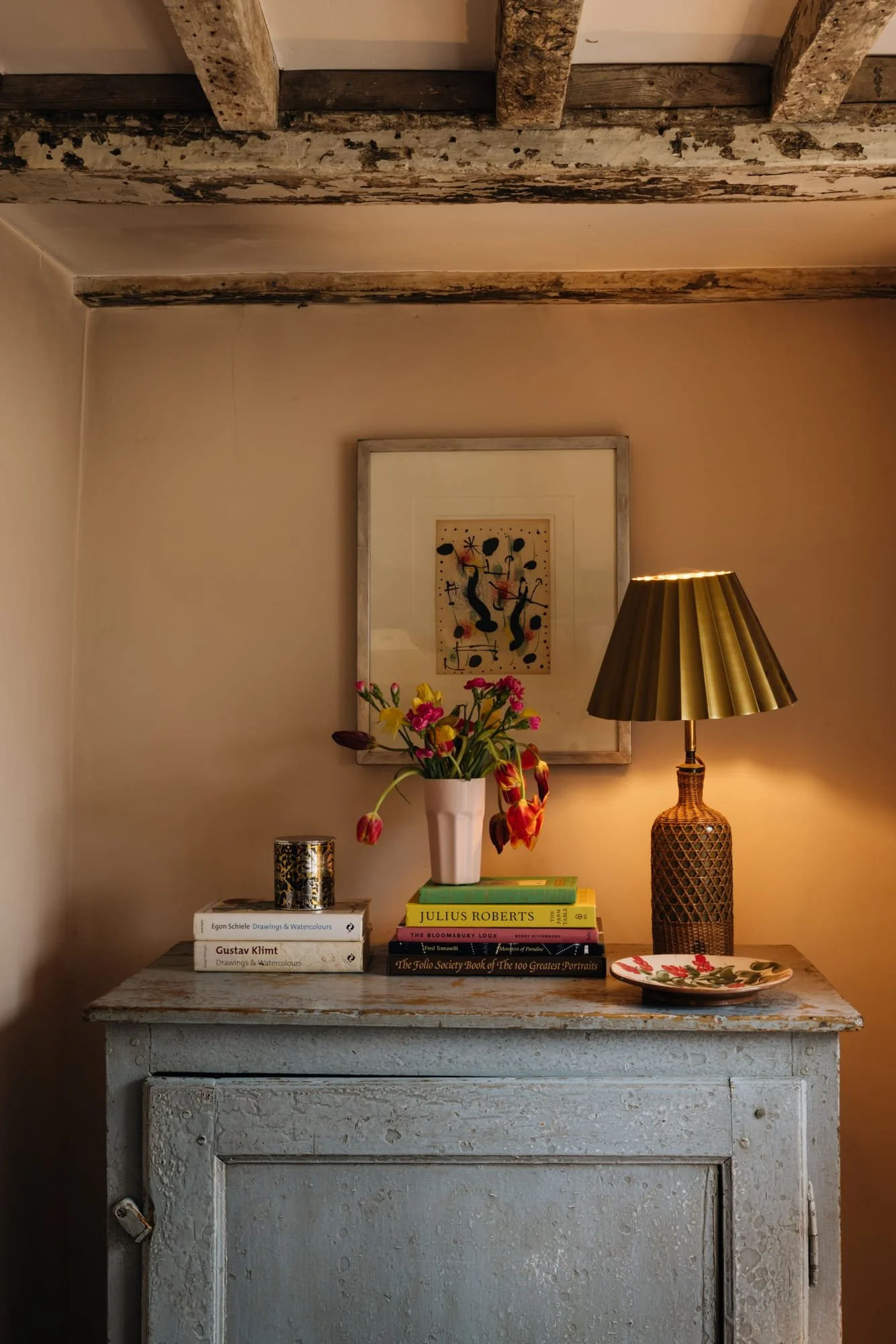 A rustic wooden sideboard against a beige wall with a framed abstract artwork, a vase of colorful flowers, a stack of books, a decorative candleholder, and a table lamp with a pleated gold shade.