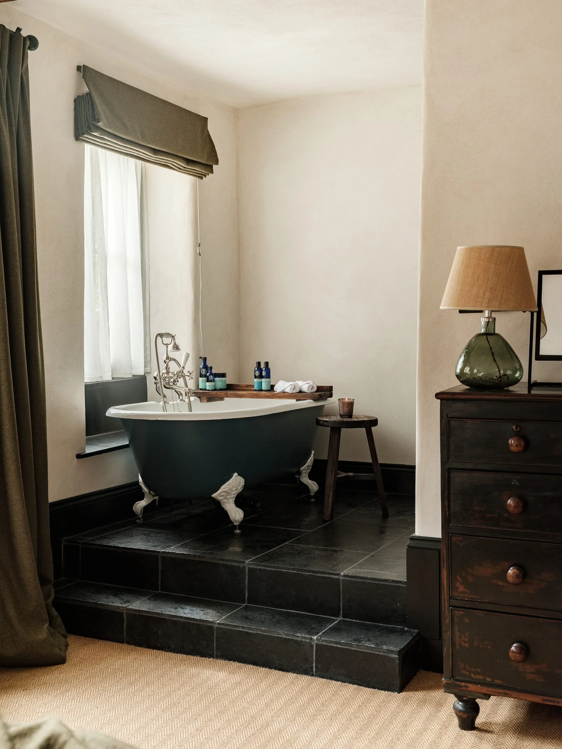 Bathroom with black clawfoot tub on raised black tiled platform, window with white curtains, small wooden side table with candle, dark wood dresser with lamp, beige carpet.
