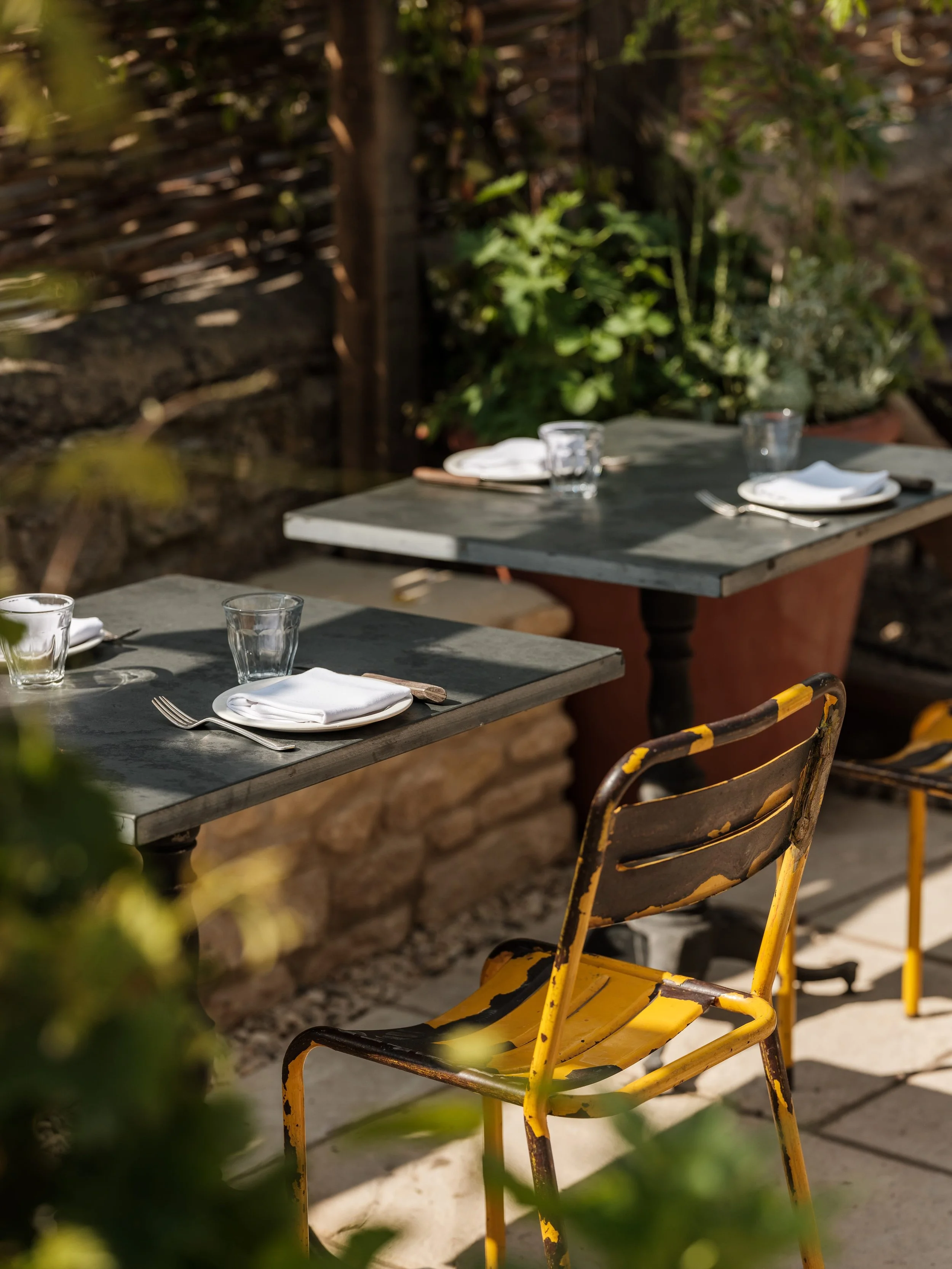 Outdoor dining setup with two tables, each with plates, napkins, glasses, and cutlery. The chairs are yellow and distressed. The setting is surrounded by greenery and a stone wall.