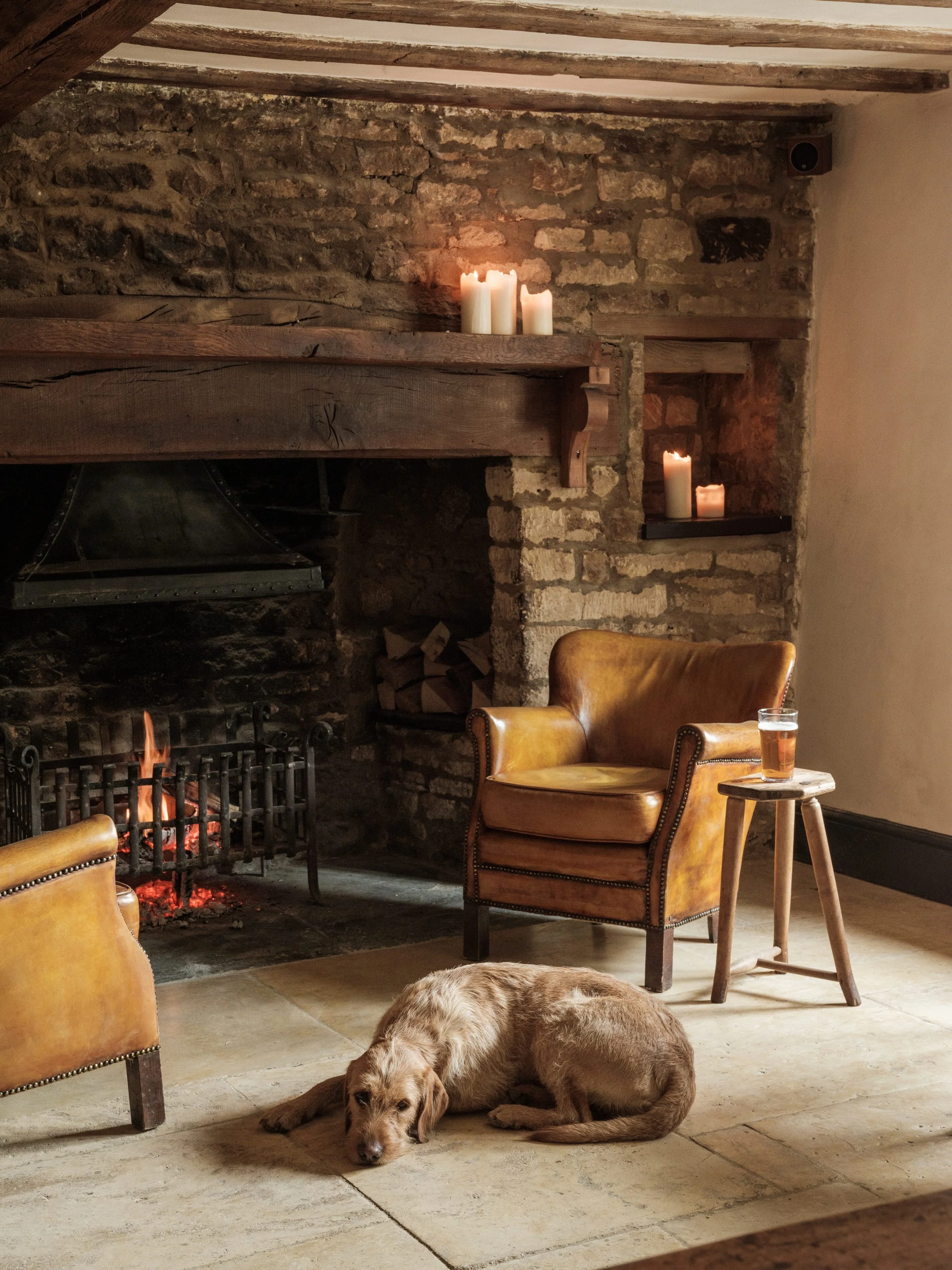 Cozy living room with a stone fireplace, wooden beams, lit candles, a leather armchair, a glass of whiskey on a small wooden table, and a dog lying on the floor.