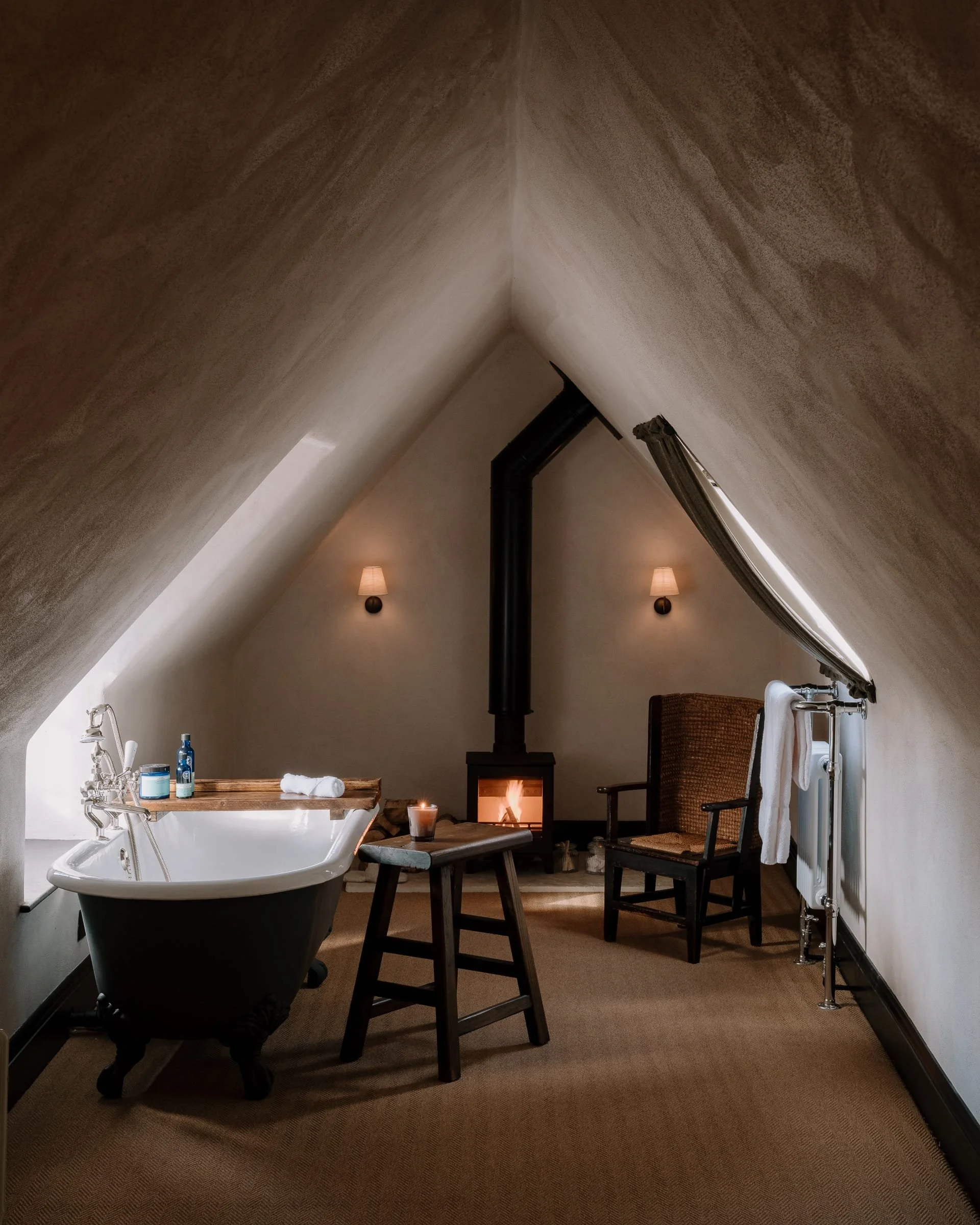 Cozy attic bathroom with a clawfoot bathtub, a wood stool with a candle, a fireplace, an armchair, and a towel rack, lit by wall sconces and natural light.