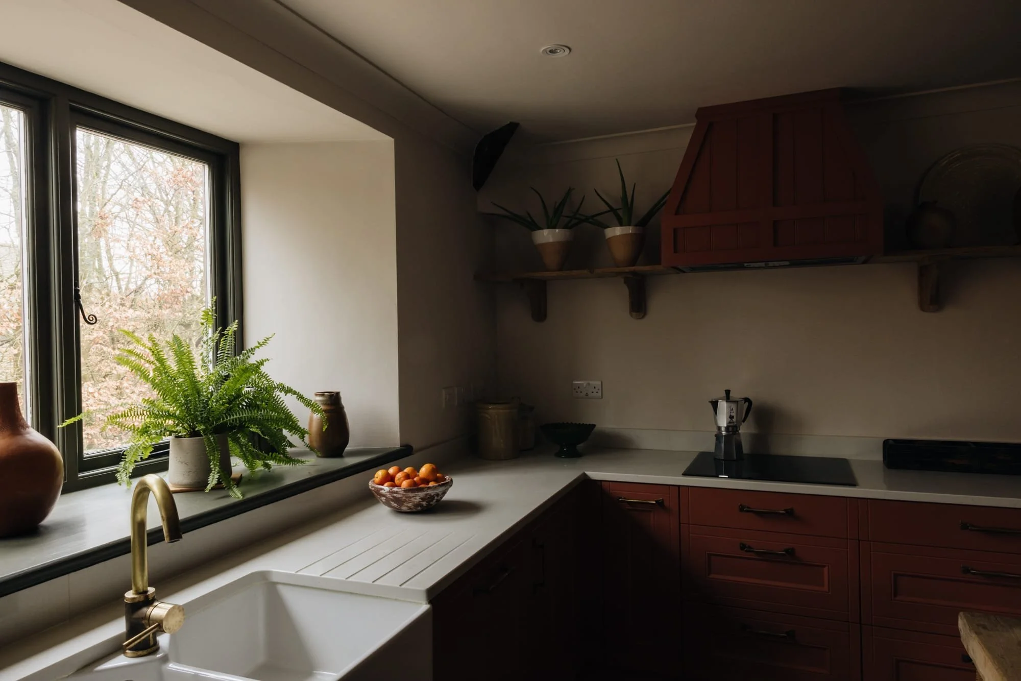 A kitchen with a window, a plant, a bowl of small orange fruits, and a brass faucet over a farmhouse sink.