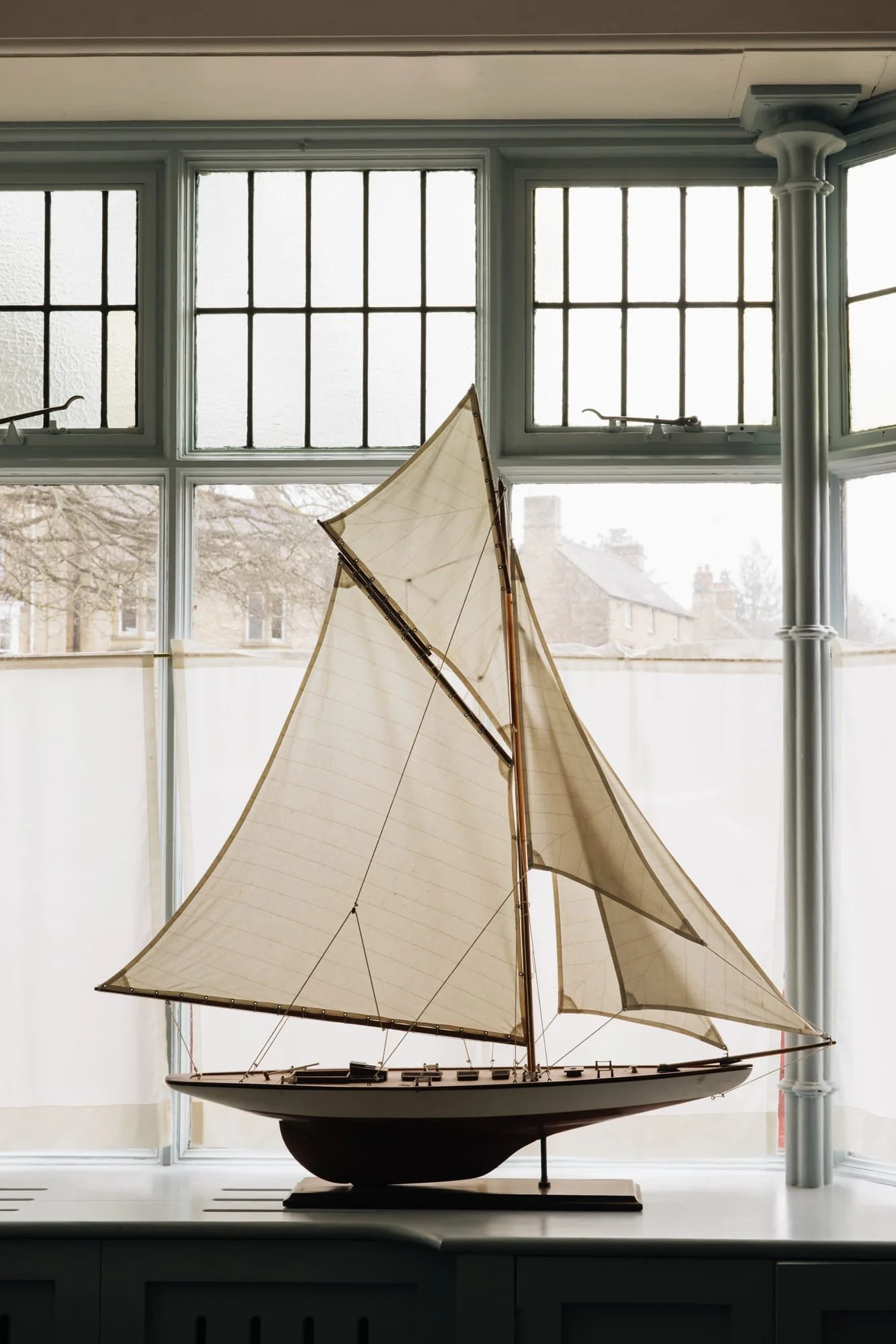 Model sailboat with white sails and wooden details placed on a windowsill in front of a large window with multiple panes.