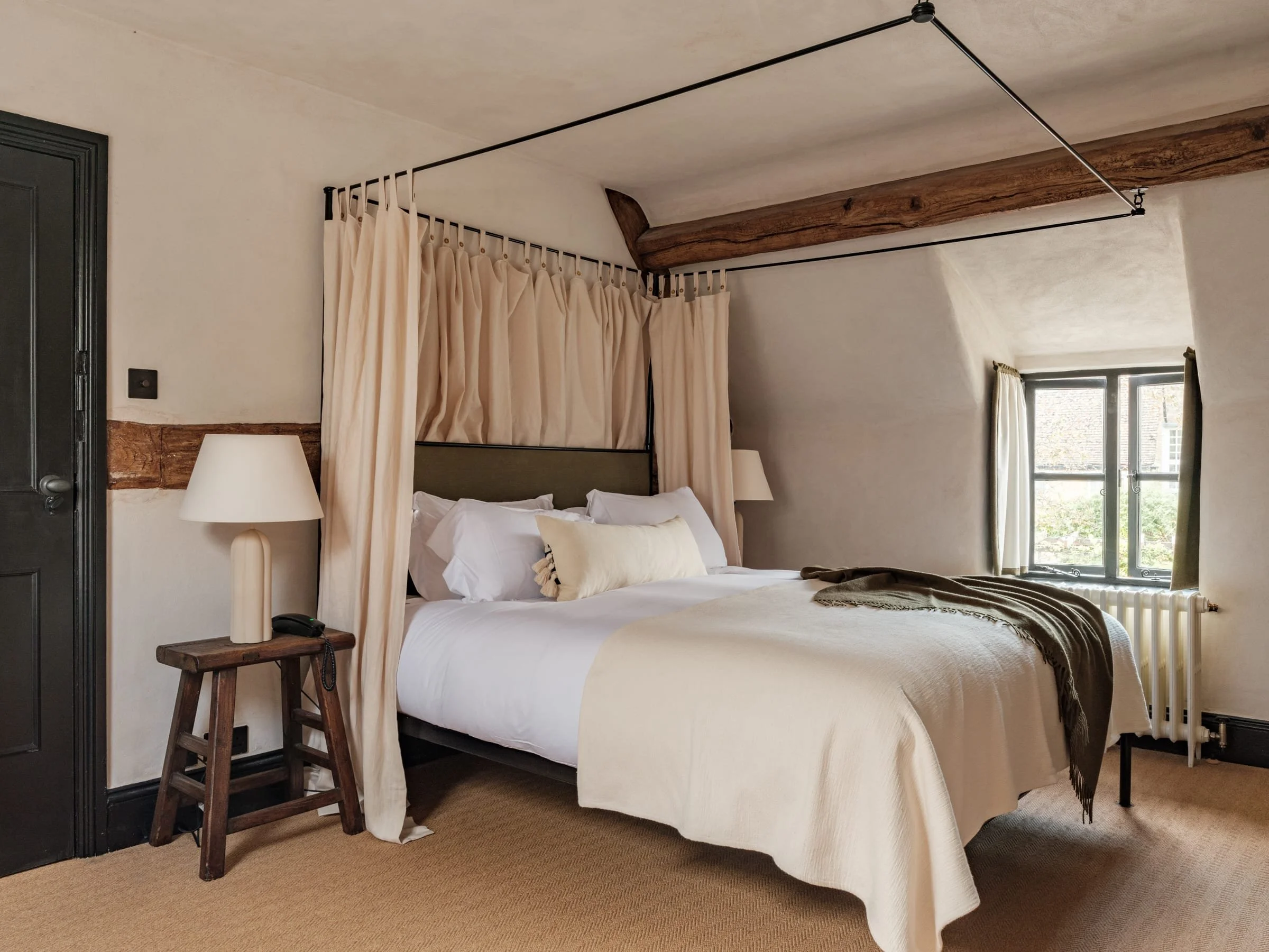 A cozy bedroom with a bed featuring white linens, multiple pillows, and a dark throw blanket. A canopy of beige curtains is suspended above the bed. There are two bedside tables, each with a lamp, and a small wooden stool beside one. The room has a s