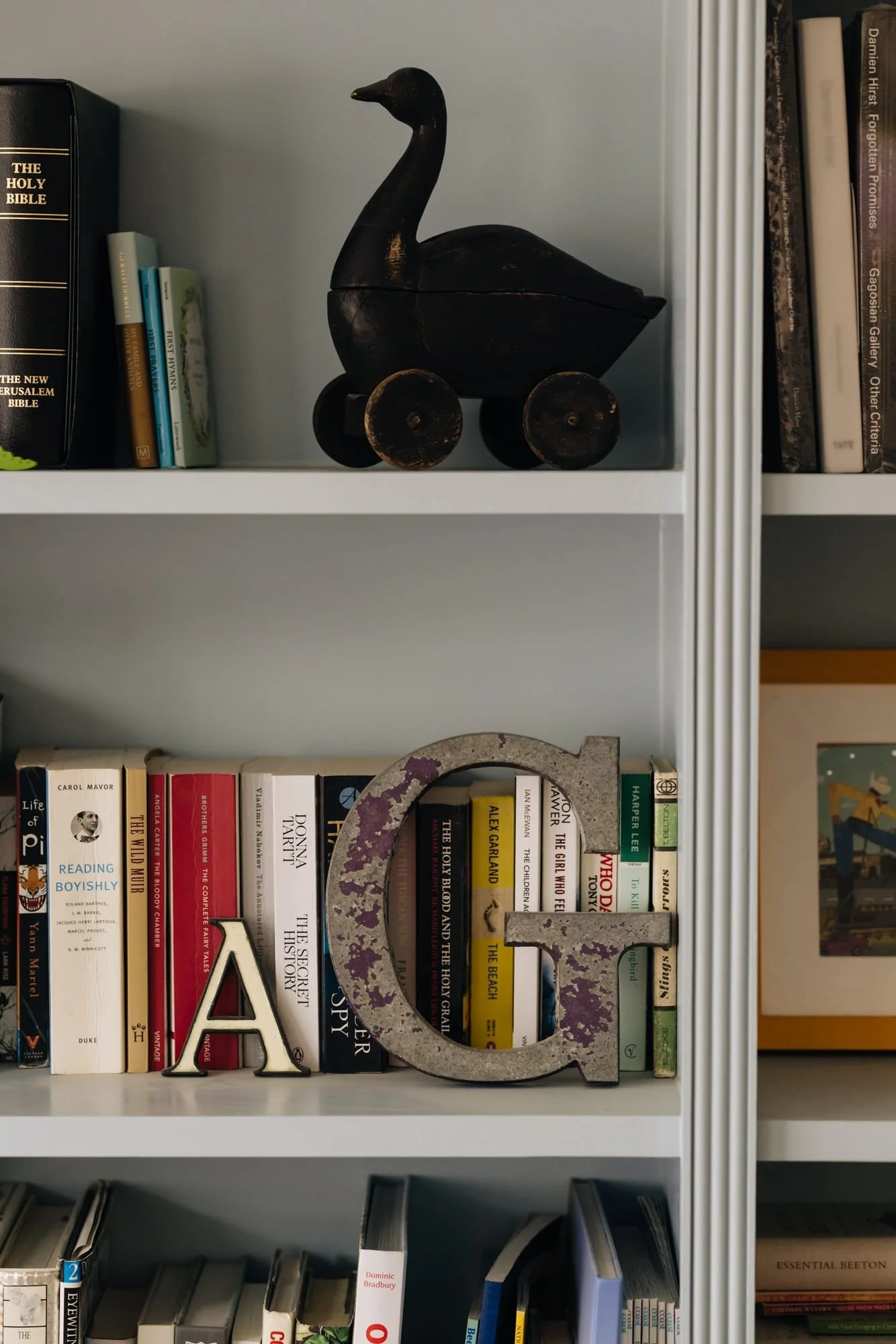 Bookshelf with a black duck-shaped wooden toy on the top shelf, and a large metal letter 'G' with signs of rust on the middle shelf, among various books.