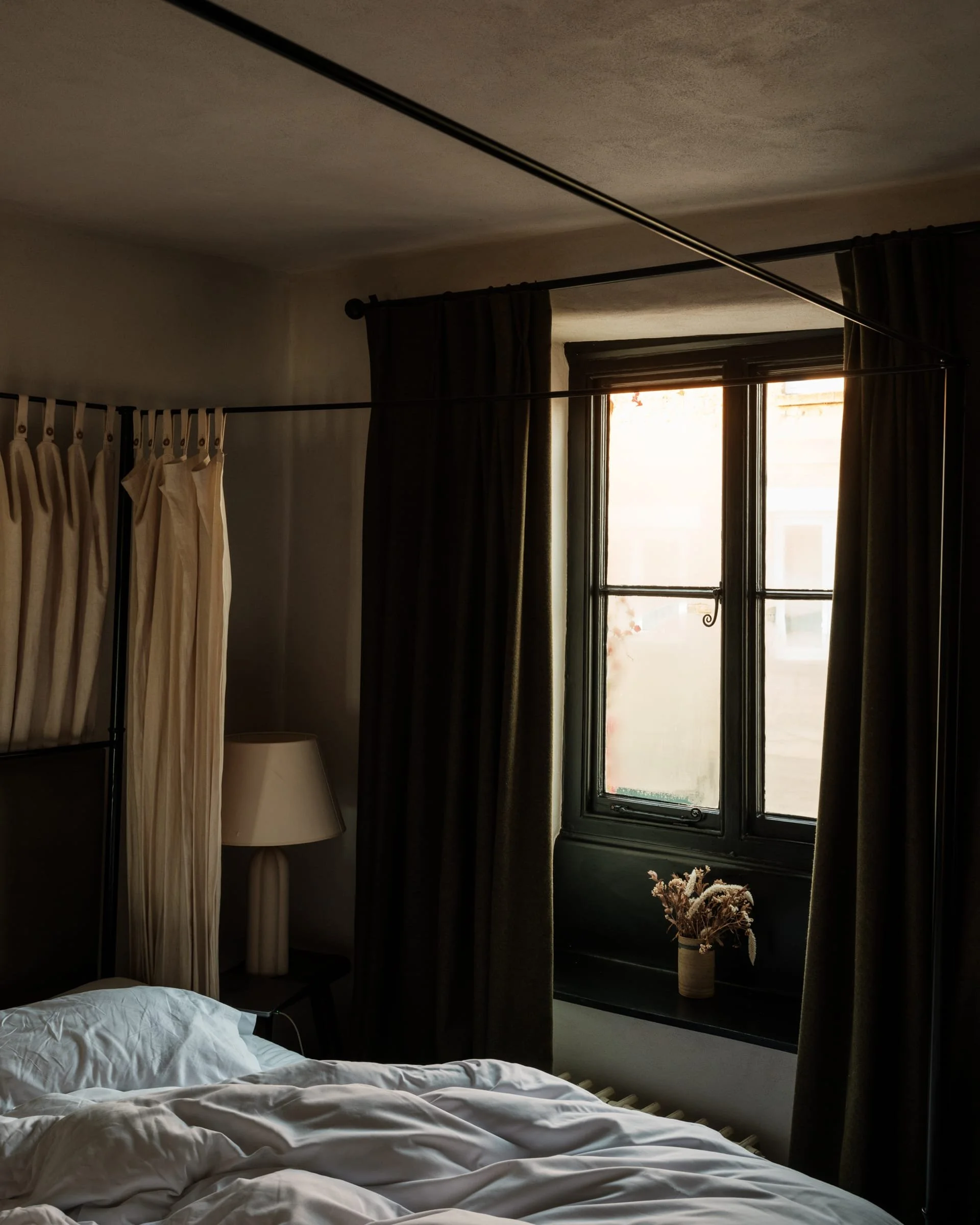 A dimly lit bedroom with a bed, a side table with a lamp, a window with black framing and dark curtains, and a bouquet of flowers in a vase on the window ledge.