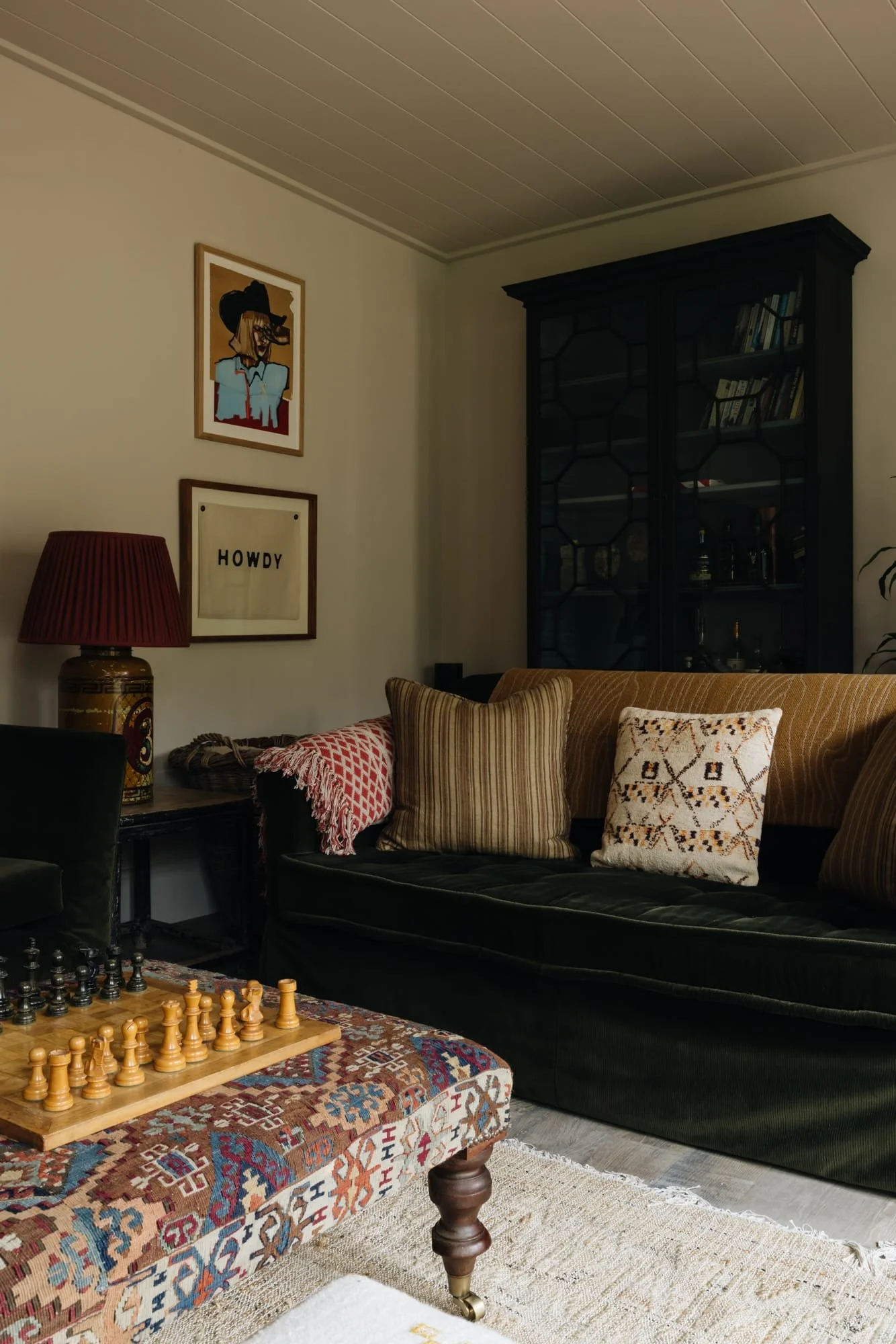 A cozy living room with a black couch, striped cushions, and a decorative pillow, a vintage style patterned ottoman with a chess set, a side table with a red lamp, framed wall art, and a black bookshelf behind the couch.