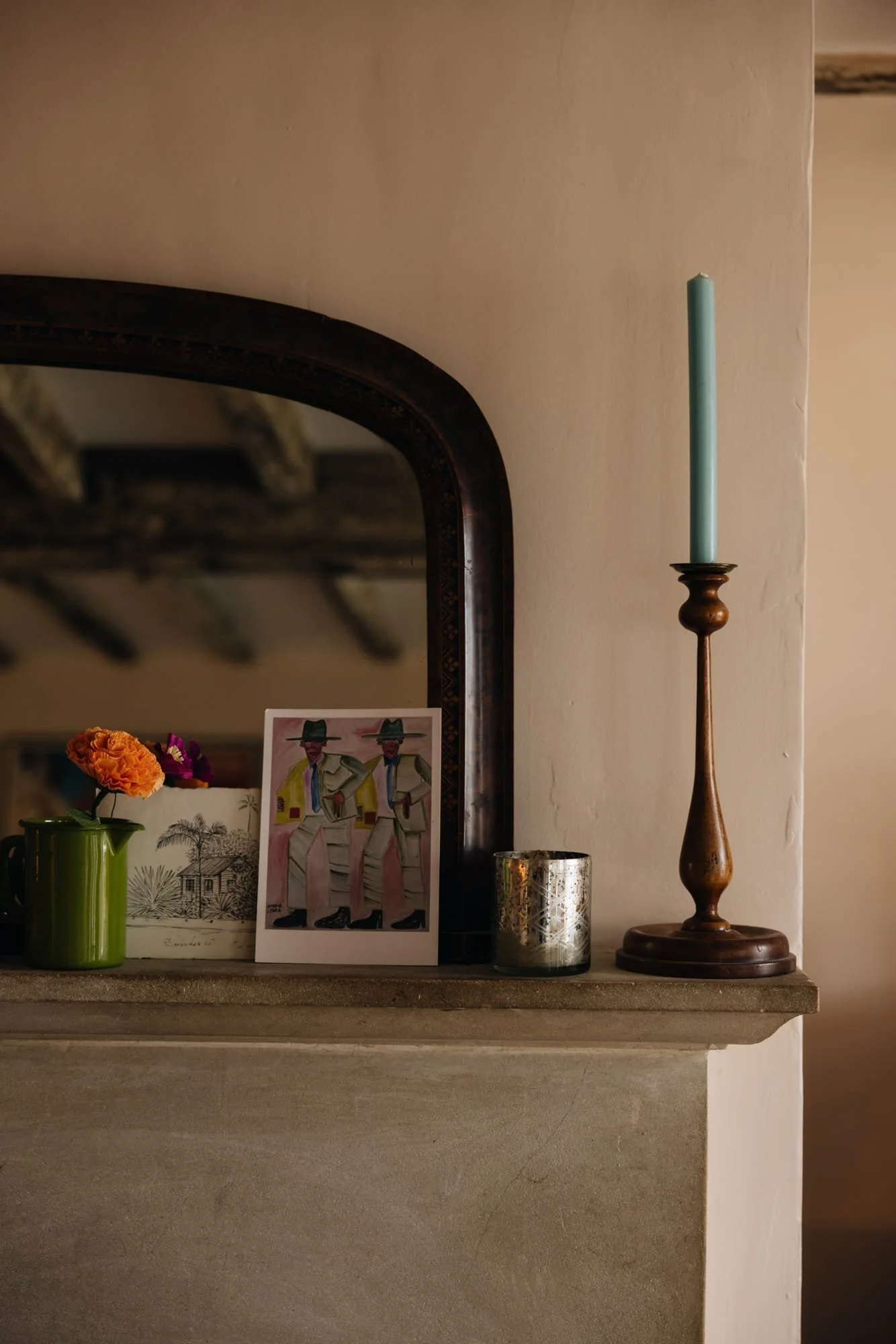 A mantelpiece with a black mirror, a green mug with orange and purple flowers, a photograph of two men in traditional Mexican attire, a metallic candle holder, and a wooden candlestick with a blue candle.