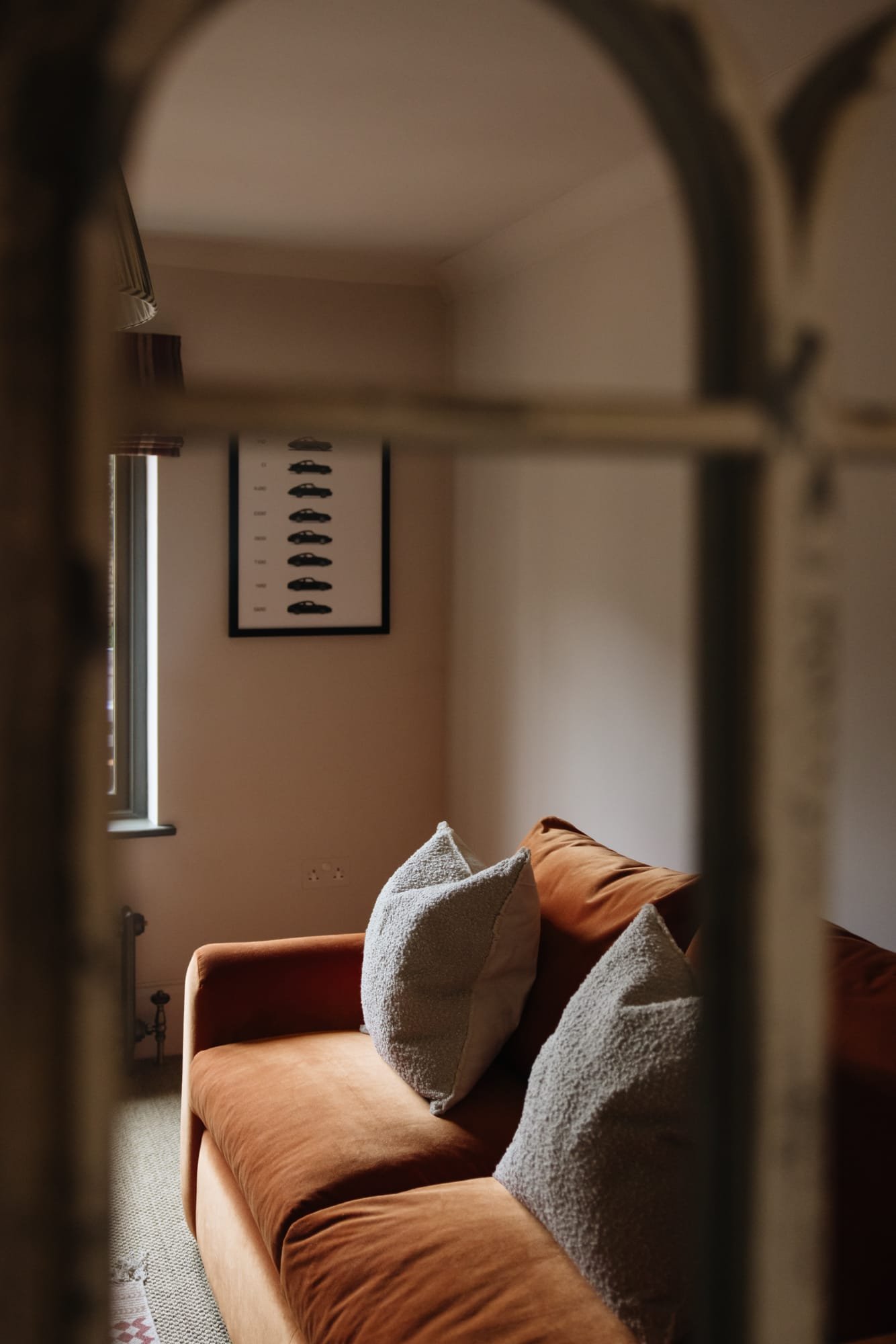 A living room viewed through a metal or wooden grid, featuring a brown velvet sofa with textured pillows and a wall hanging photo of shoes in the background.