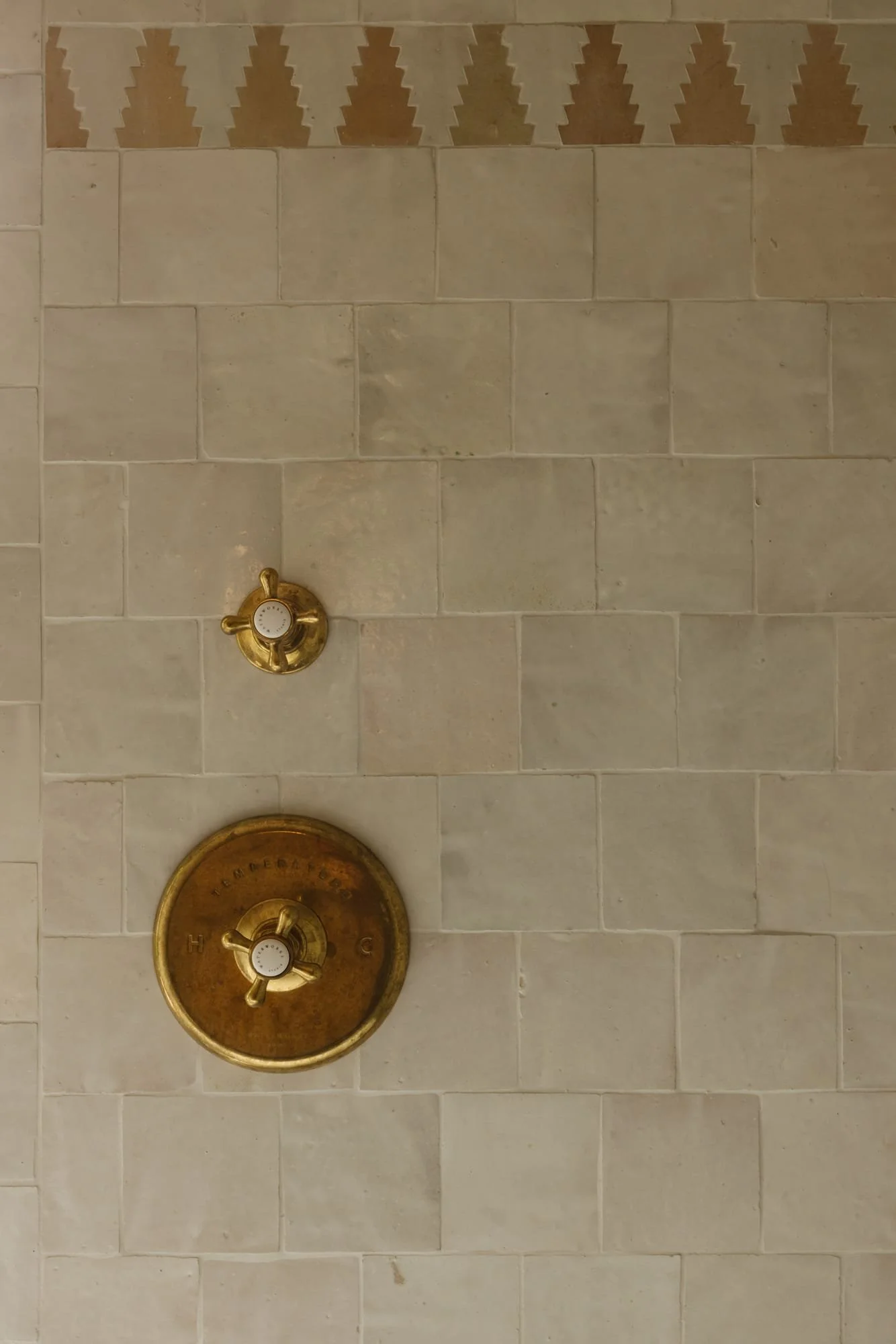 Close-up of a shower wall with beige tiles, featuring a brass shower control valve and a matching brass on/off knob, with tiles patterned at the top.