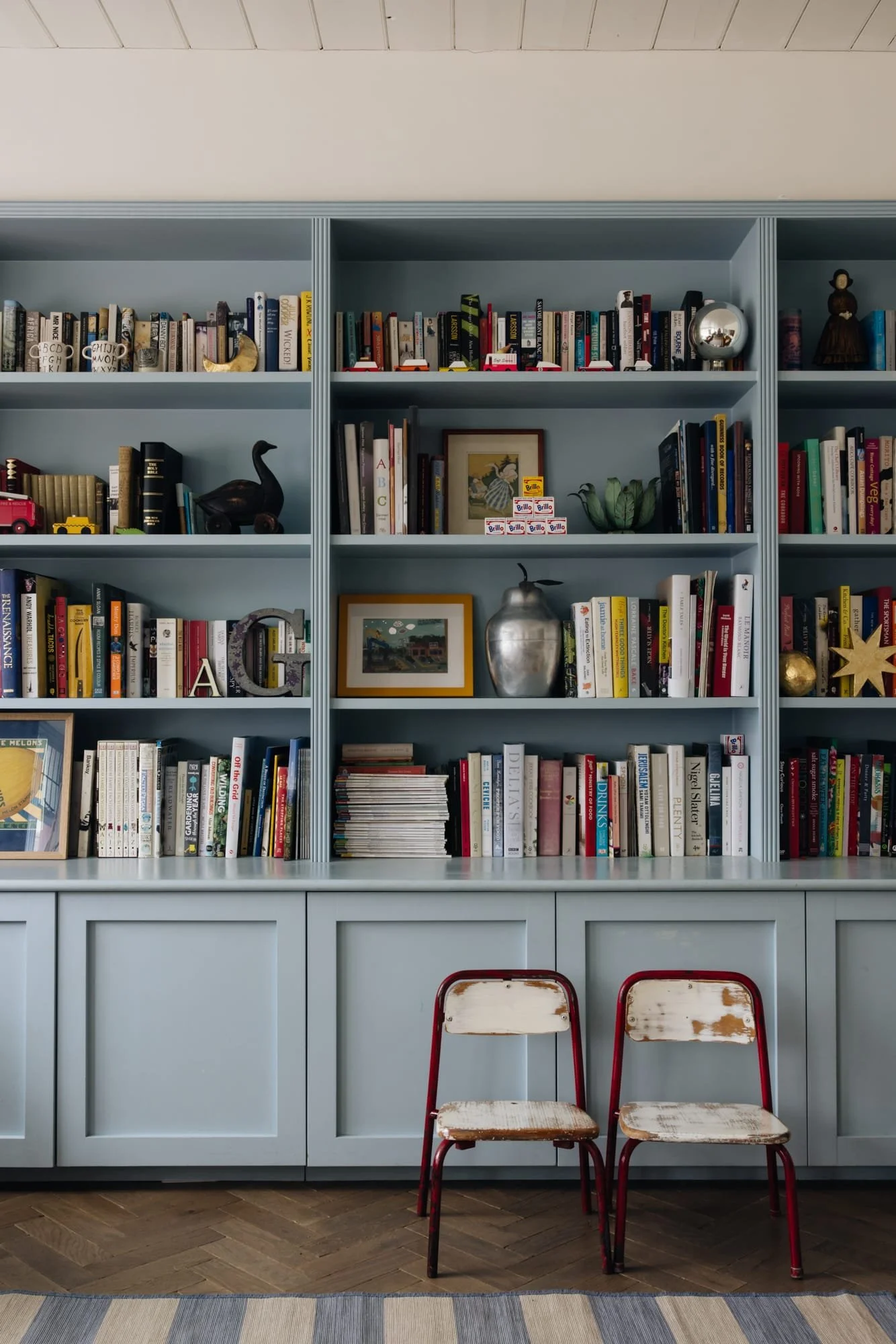 A light blue bookshelf filled with books, decorative objects, and framed pictures, with two worn wooden chairs with red frames in front of it.
