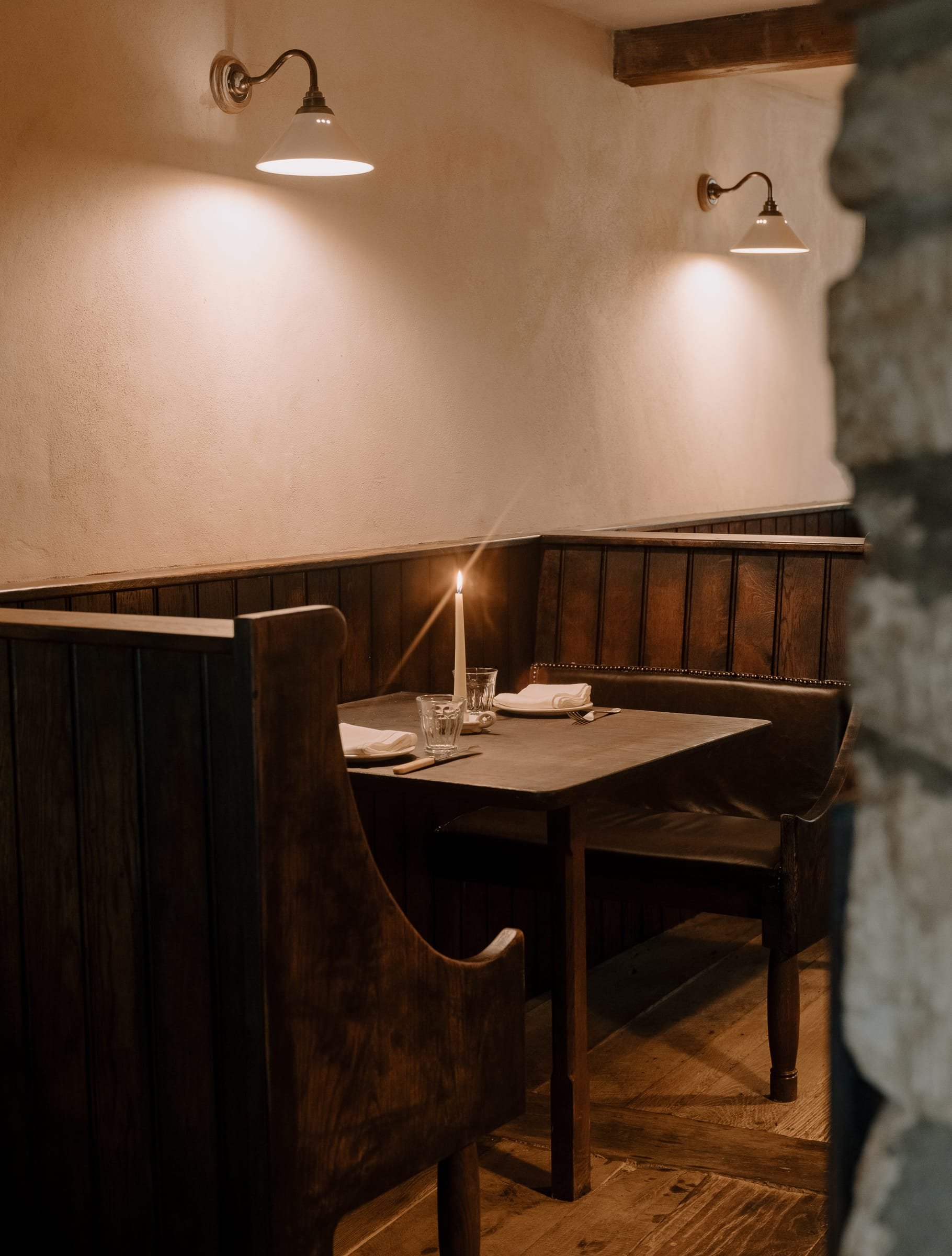 A corner of a cozy, dimly lit restaurant booth with a table set with a candle, glasses, and napkins, featuring dark wood paneling and warm wall-mounted lights.