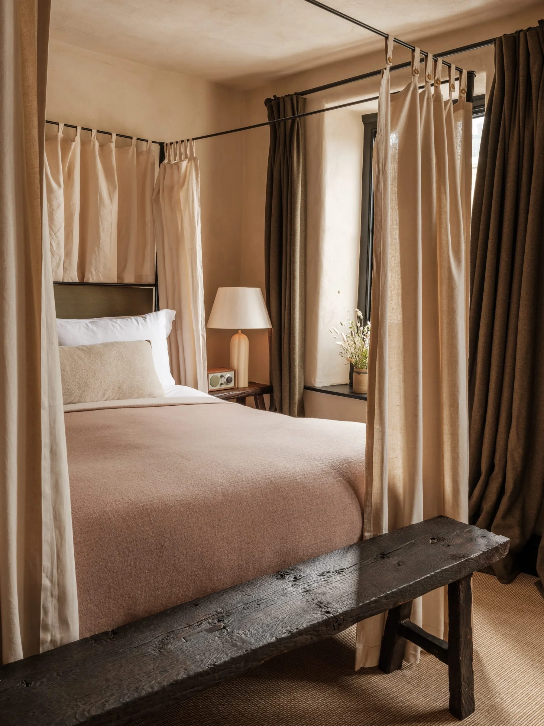 A cozy bedroom with a platform bed, beige bedding, and a four-poster canopy with curtains. There's a wooden side table with a white lamp and window with curtains and a small plant on the windowsill.