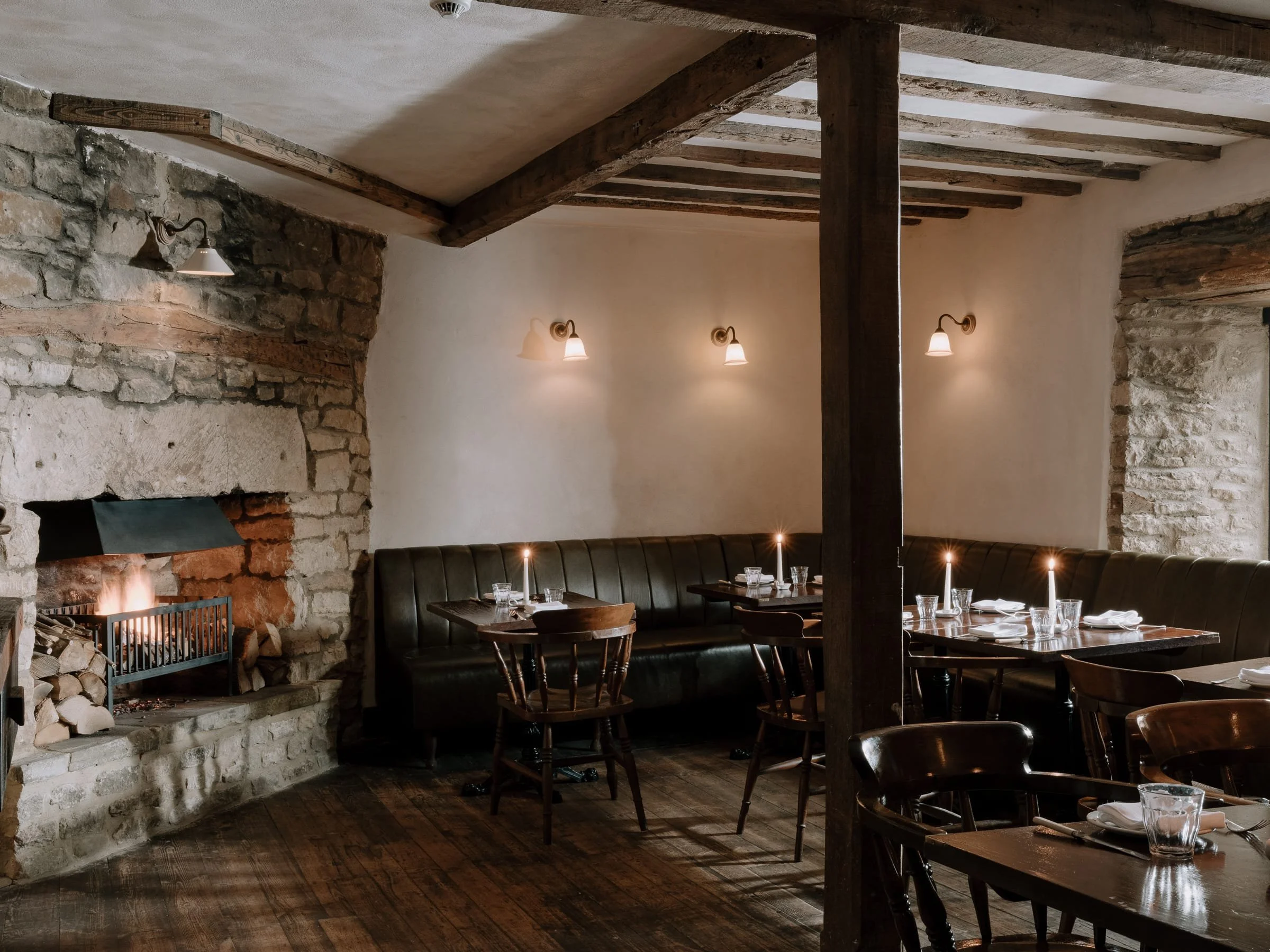 Cozy dining room with stone and wooden walls, a fireplace, wooden beams, dark wood chairs, a black leather bench, and candlelit tables.