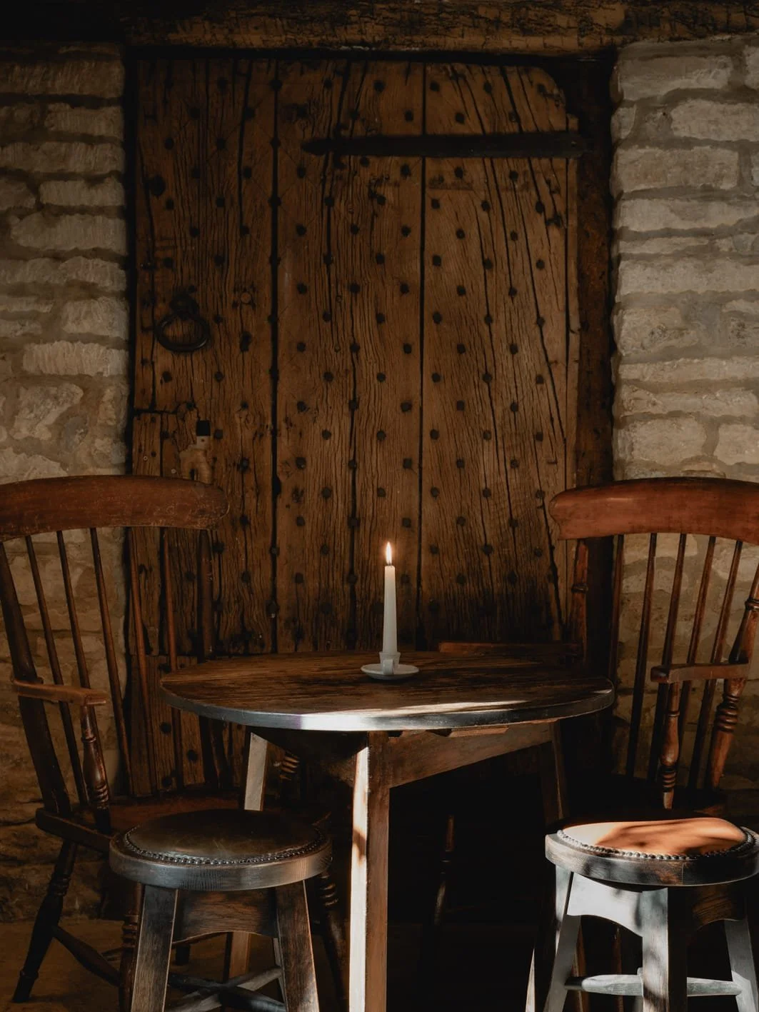 A rustic dimly lit room with a small round wooden table, two chairs, a candle, and a large wooden door with black metal hardware, surrounded by stone walls.