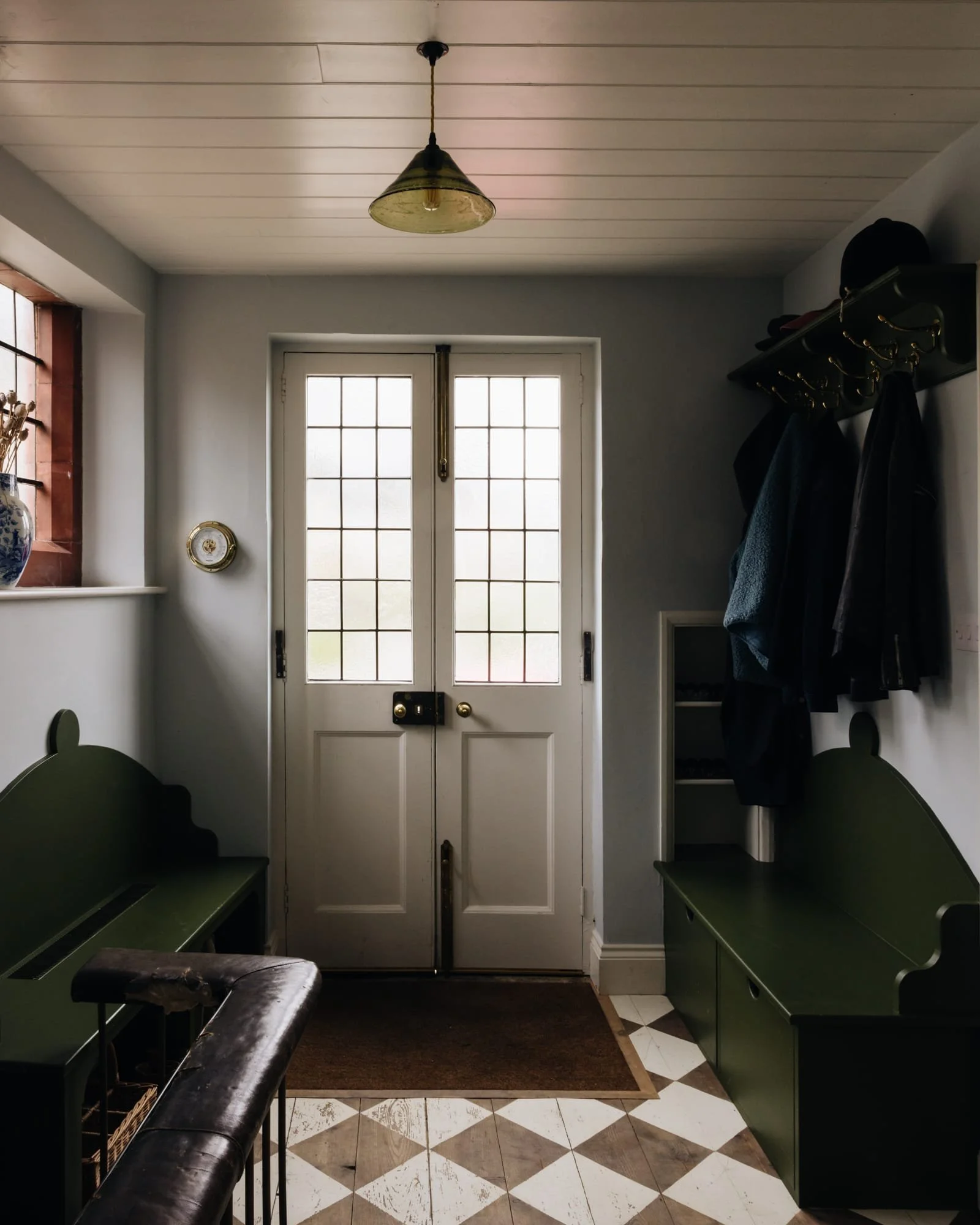 Entryway with white double doors with glass panes, green benches on each side, coat rack with jackets, window with a vase on the sill, and a ceiling light fixture.