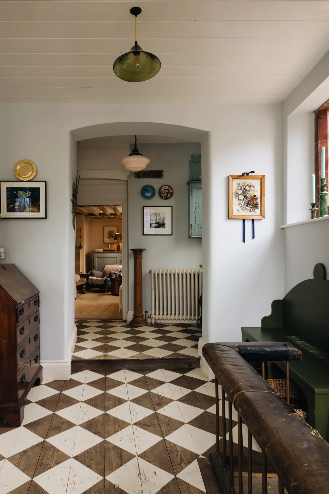 Interior view of a vintage style house with checkered floor tiles, a staircase with a wooden banister, framed artwork on the walls, and a glimpse into a cozy living room with armchairs and lamps.