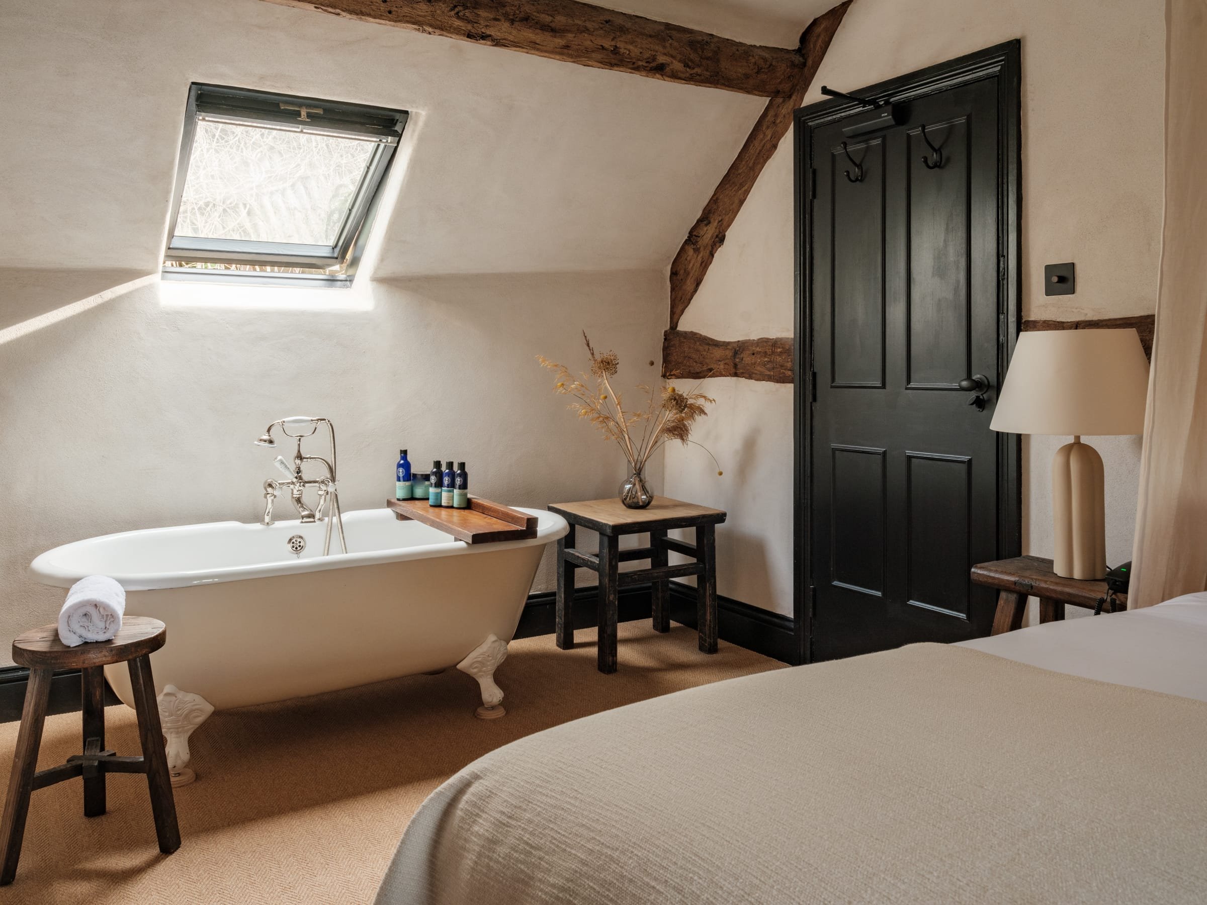 A cozy bedroom with a bathtub, a skylight window, a small wooden side table with rolled towel, a black wardrobe, a bedside table with a lamp, and decorative dried plants in a vase.