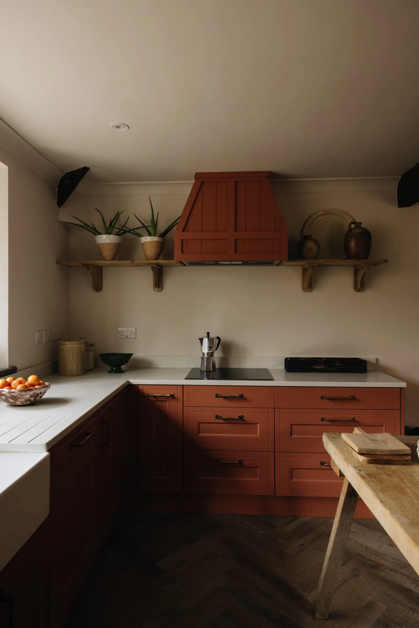 Kitchen with red cabinets, open shelving with potted plants and decorative vases, stove with moka pot, wooden table with cutting boards, bowl of oranges, and dark wood flooring.