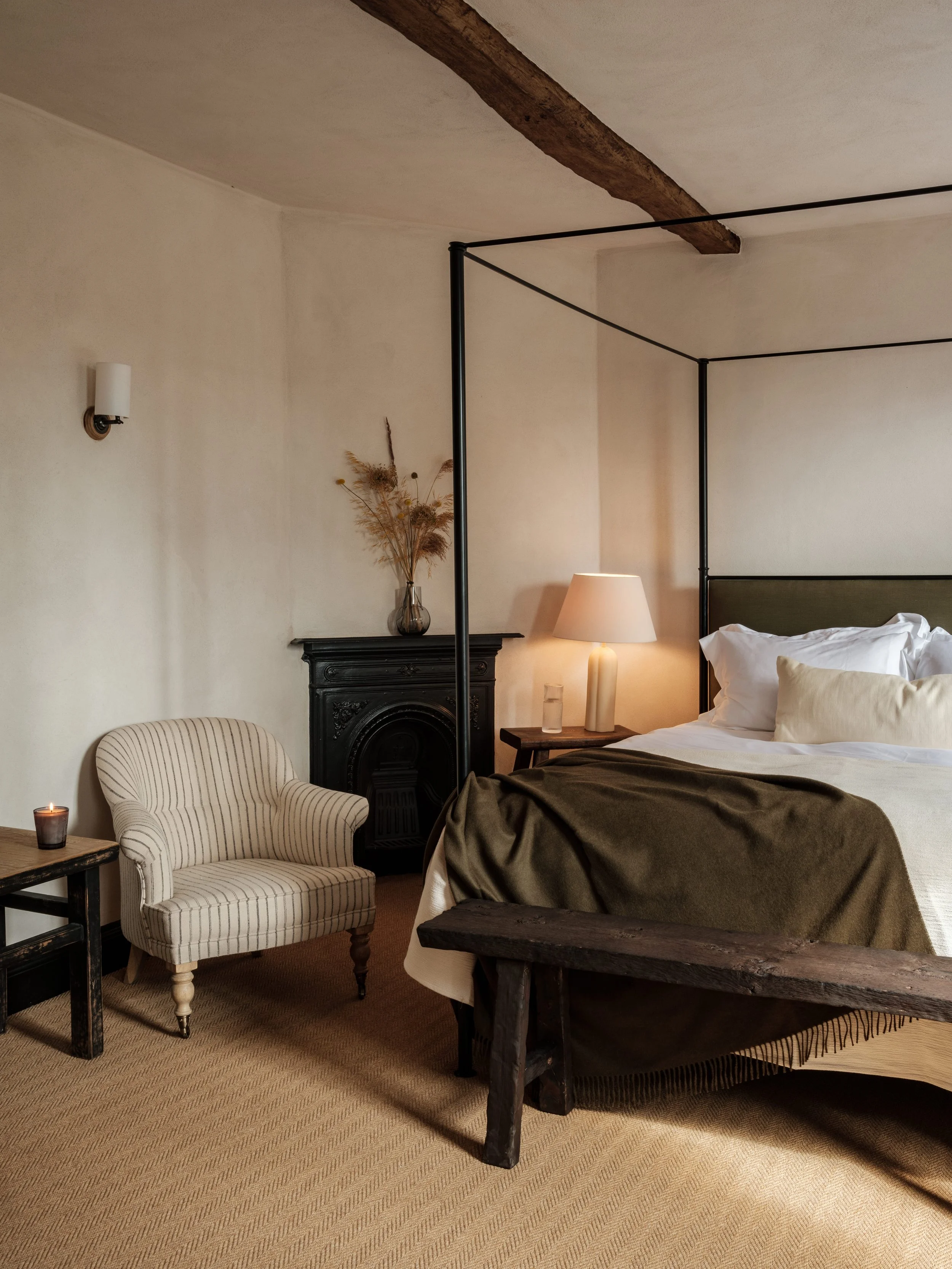 A cozy bedroom with a beige carpet, a bed with white linens and brown blanket, a wooden bench at the foot, a side table with a lamp, an armchair, and a decorative vase with dried plants on a black fireplace mantel.