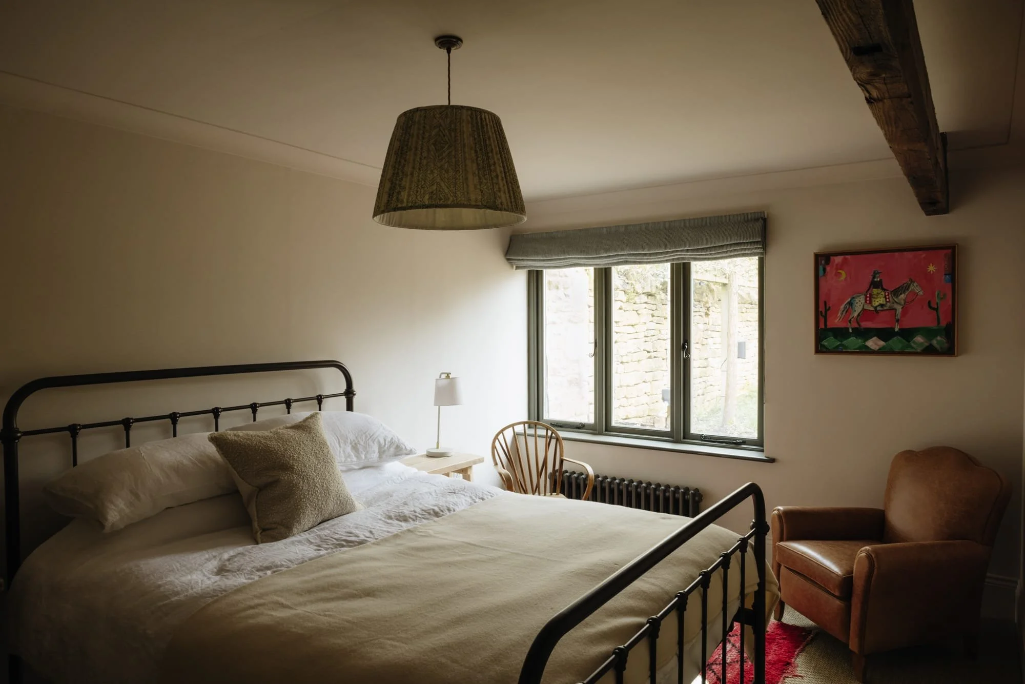 A cozy bedroom with a metal-framed bed, beige bedding, a wooden nightstand with a small lamp, a wooden chair by the window, and a brown armchair near a colorful wall art piece depicting a horse on a pink background, with a large window showing an out
